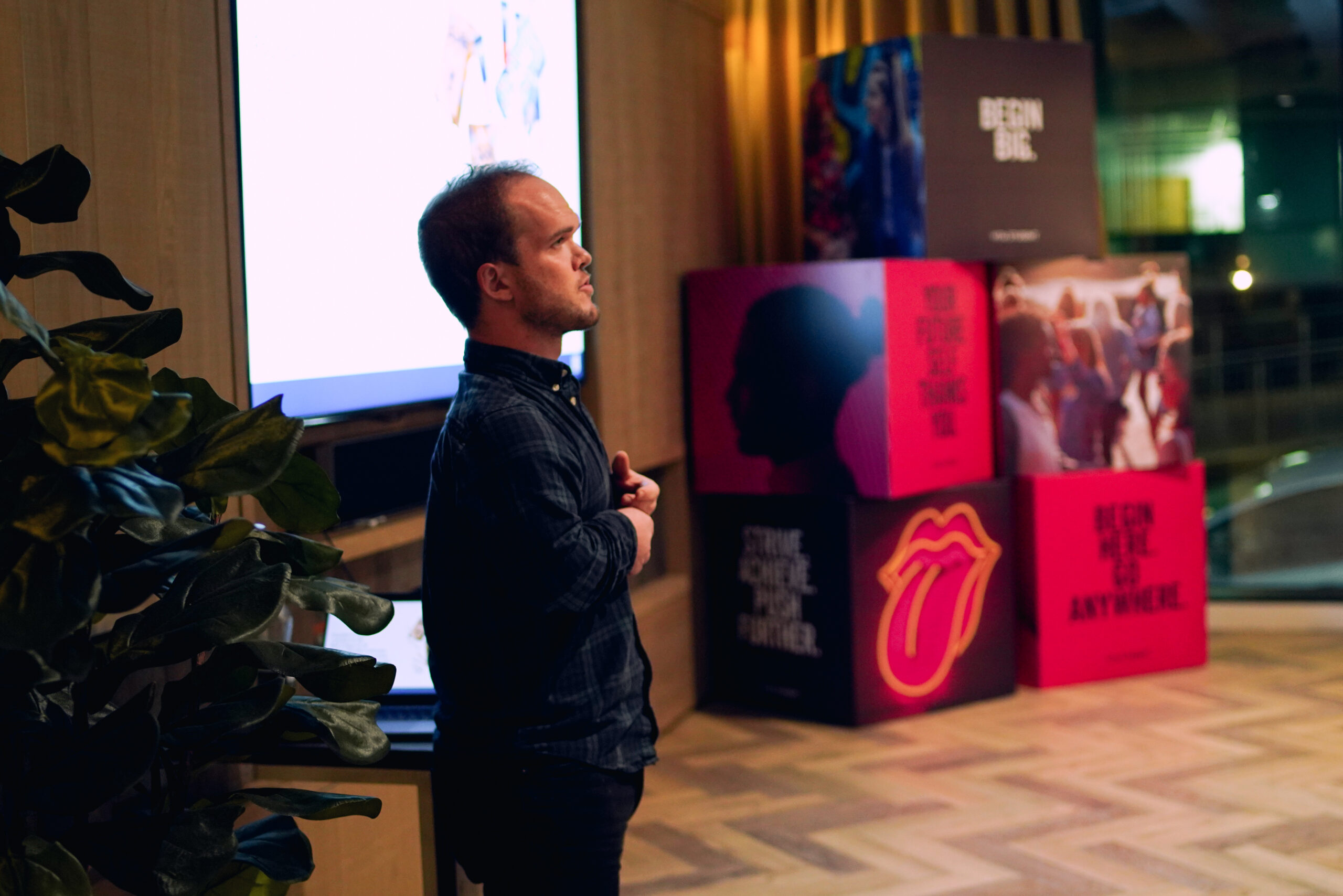 A man gives a lively presentation in a room with wood flooring and vibrant plants, his enthusiasm reflecting the colorful stacked boxes adorned with graphics, including the striking Vita tongue logo projected on the screen.