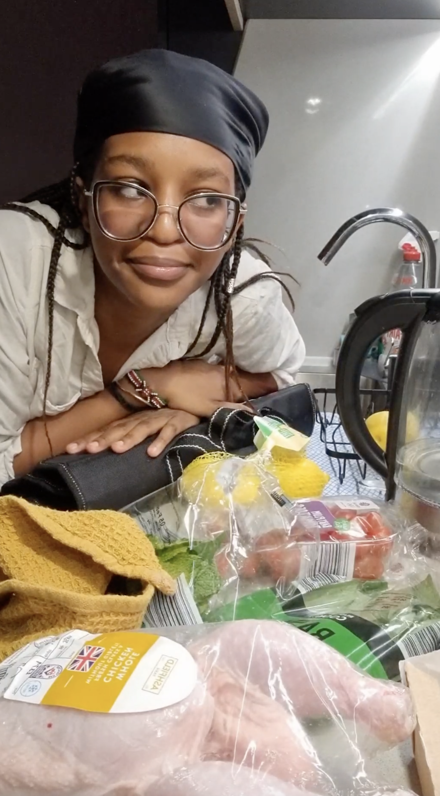 A person with glasses and a headscarf leans on a counter, surrounded by groceries including a bag of lemons, tomatoes, and leafy greens. A yellow cloth and a kettle are also visible in the kitchen setting.