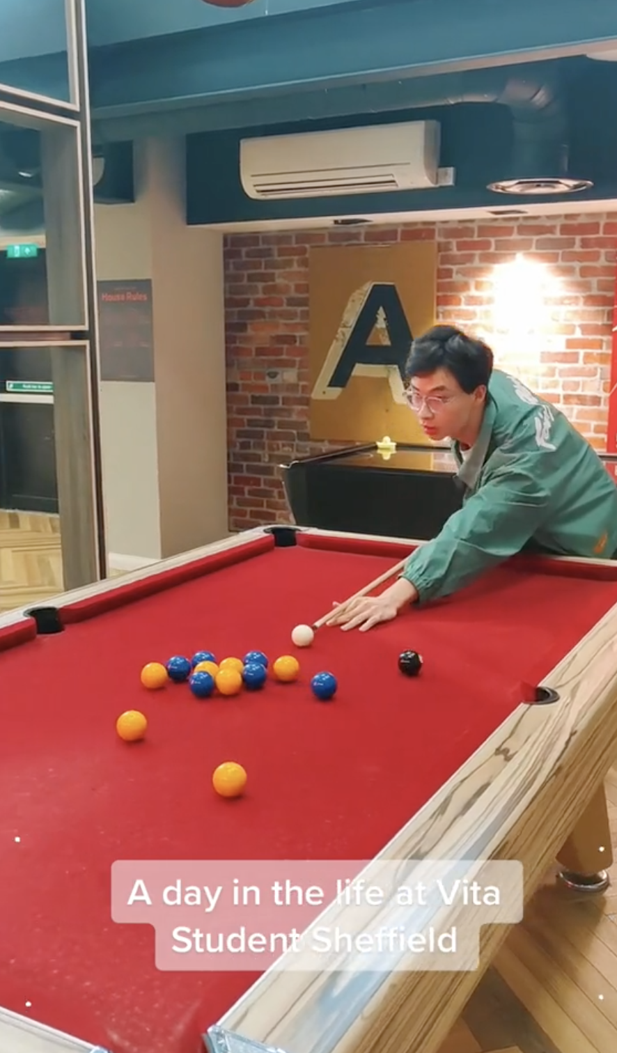 A person playing pool in a game room with a red-felted table and colorful balls. There is a brick wall and a yellow and black letter "A" sign in the background. Text on the image reads, "A day in the life at Vita Student Sheffield.