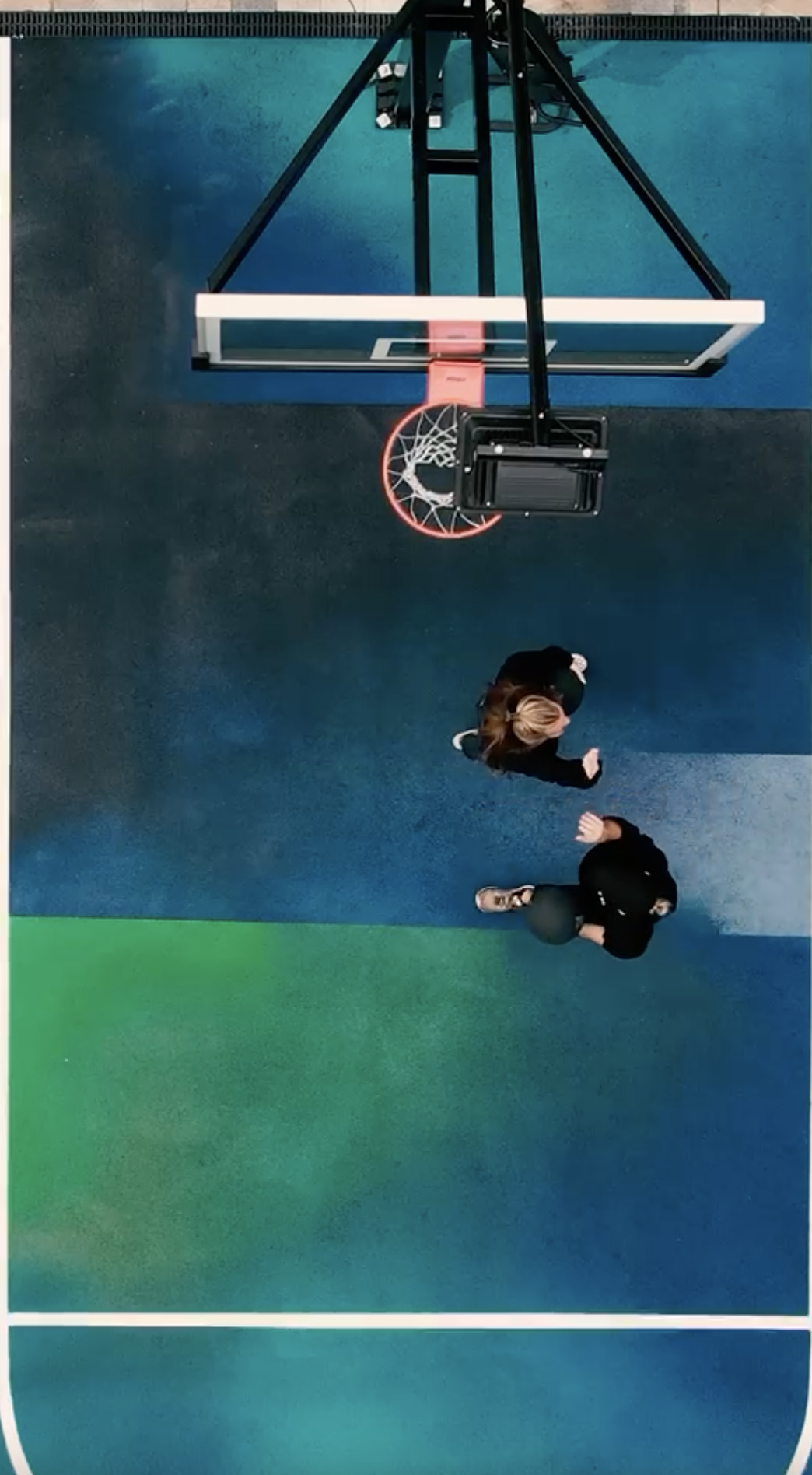 Overhead view of a basketball court with two people walking near the hoop. The court has blue, green, and black sections. The basketball hoop casts a shadow on the ground.