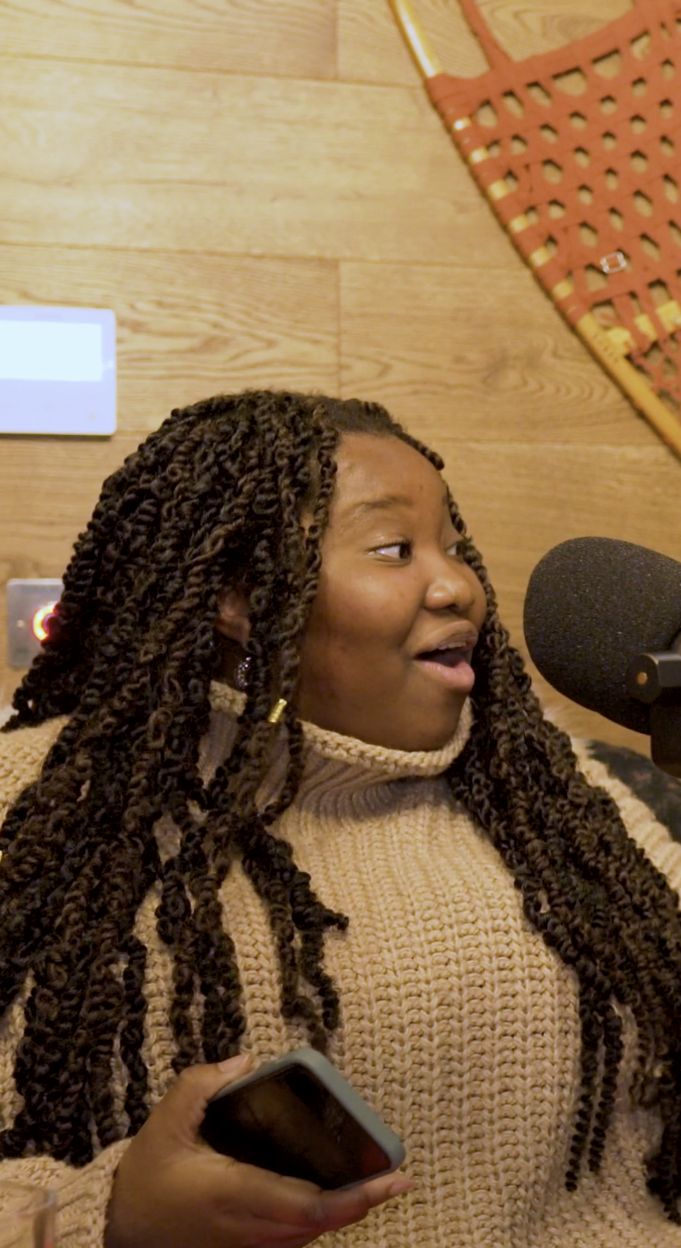 A person with long braided hair is speaking into a microphone while wearing a beige sweater. The background features a wooden wall with a snowshoe hanging on it.