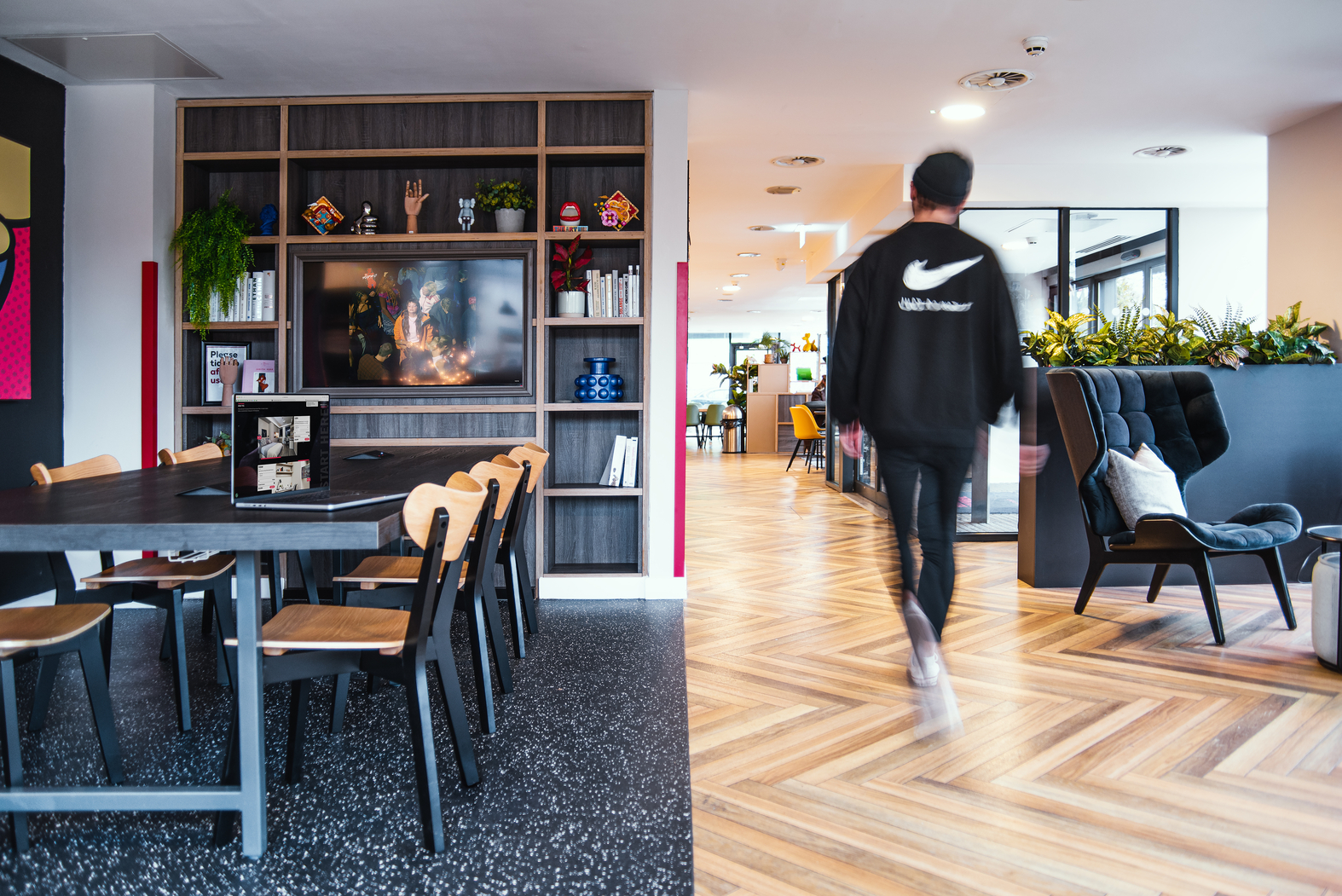 A modern office space with wooden flooring and contemporary furniture evokes the feel of upscale student accommodation. A blurred figure walks past a table with chairs, while shelves filled with books and decor line one wall, complemented by plants and minimalist art.