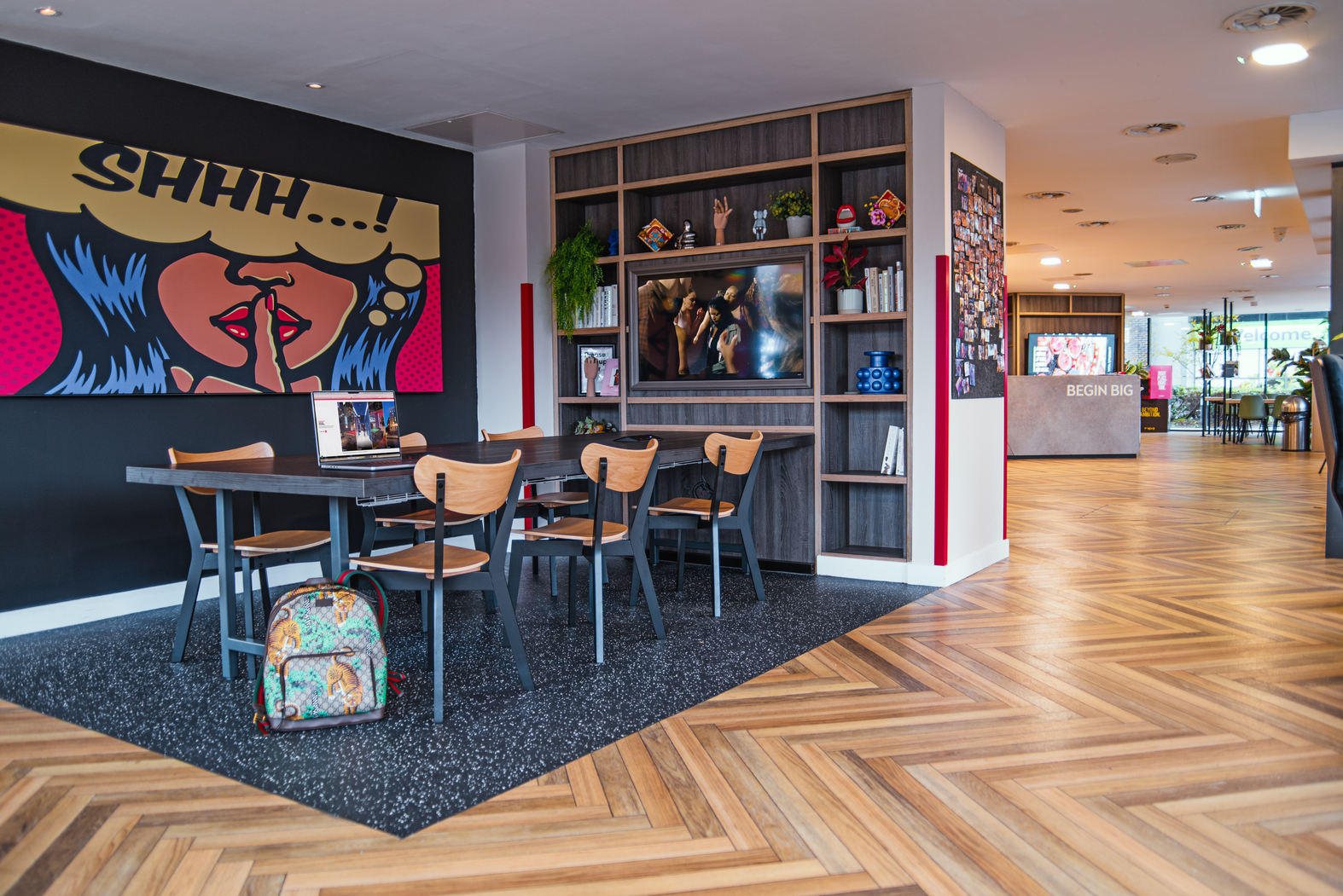 A modern, open-plan office space with wooden herringbone flooring seamlessly blends into a student accommodation vibe. A table with chairs and a colorful backpack occupies the foreground. The back wall showcases a vibrant pop art mural, while bookshelves display decorative items and plants.