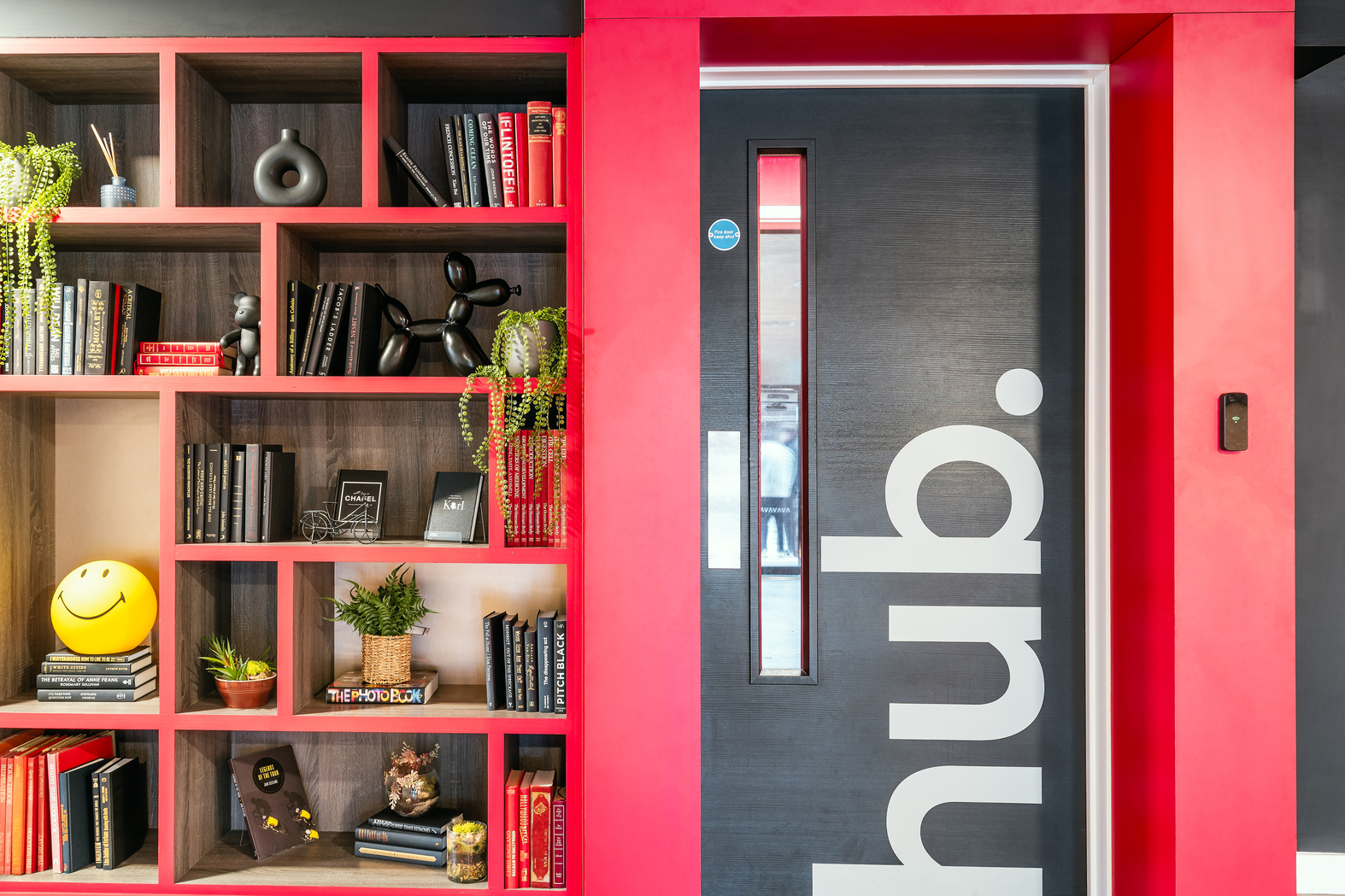 A vibrant office space reminiscent of modern student accommodation features a red bookshelf filled with books, plants, and decorative items. To the right, a dark door marked "hub" in bold white letters is framed by a red border, creating an inviting atmosphere.