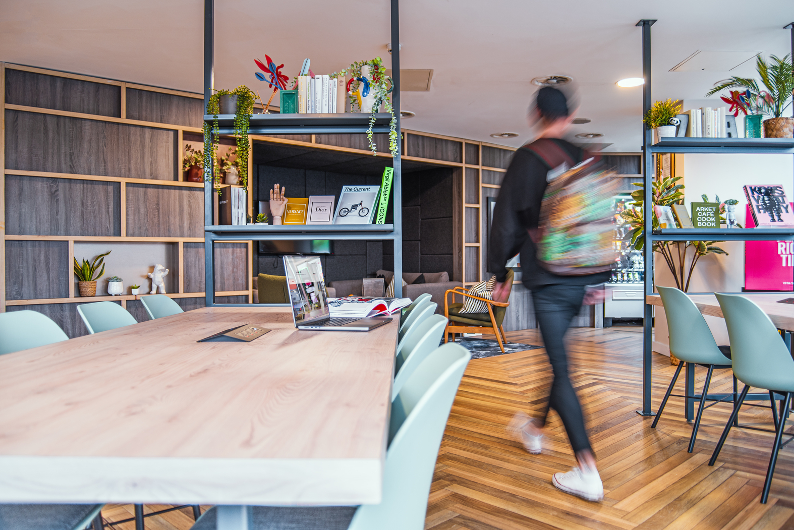 A person with a colorful backpack strolls through a modern coworking space, reminiscent of vibrant student accommodation. The area features wooden tables, blue chairs, and shelves adorned with books and plants, while an open laptop sits on one table amid charming decorative items.