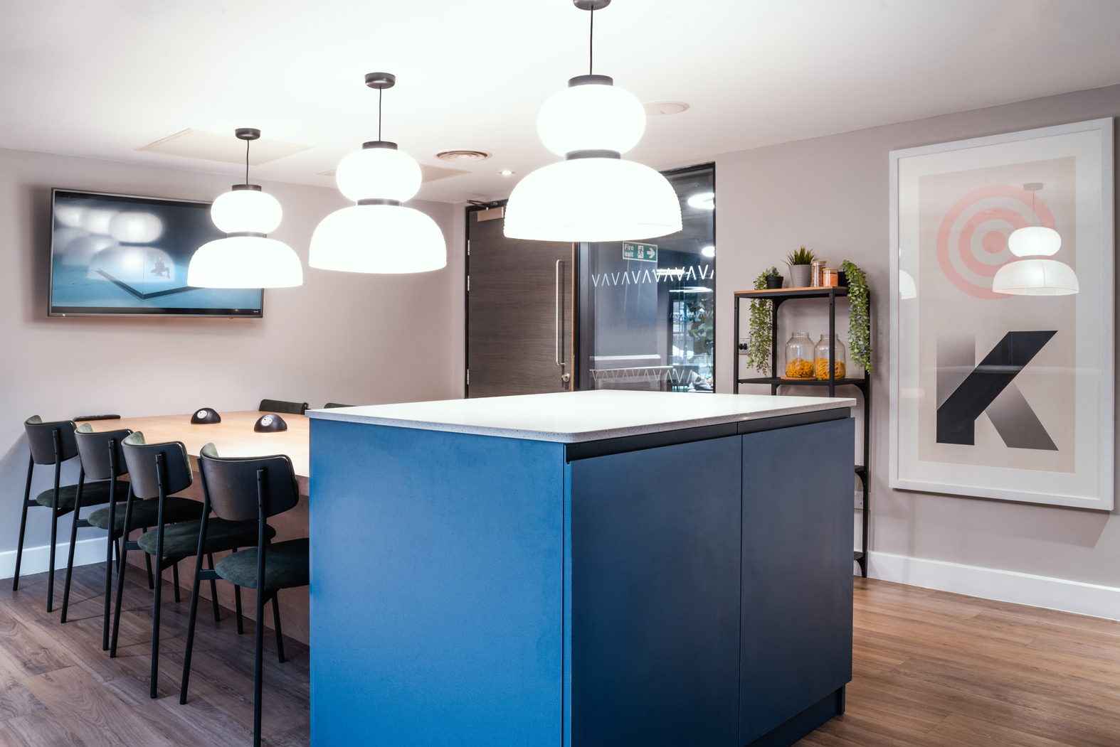 Modern conference room in a student accommodation, featuring a long wooden table surrounded by black chairs, a blue cabinet, pendant lights, a wall-mounted TV, and a framed abstract art piece. A small shelf with decorative items is near the glass doors.