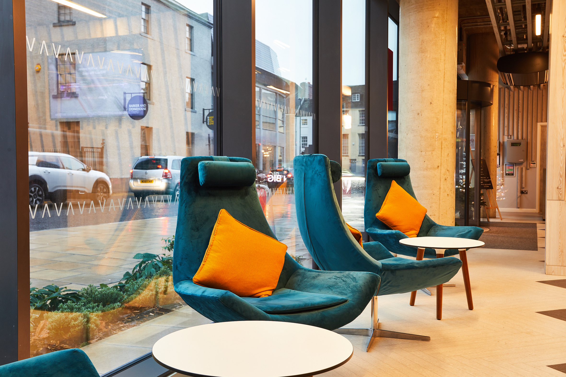 A modern seating area with teal armchairs and bright orange cushions embraces the place, facing floor-to-ceiling windows. Small round tables sit in front of the chairs. Beyond the glass, cars and buildings add a bustling backdrop to this cozy retreat.