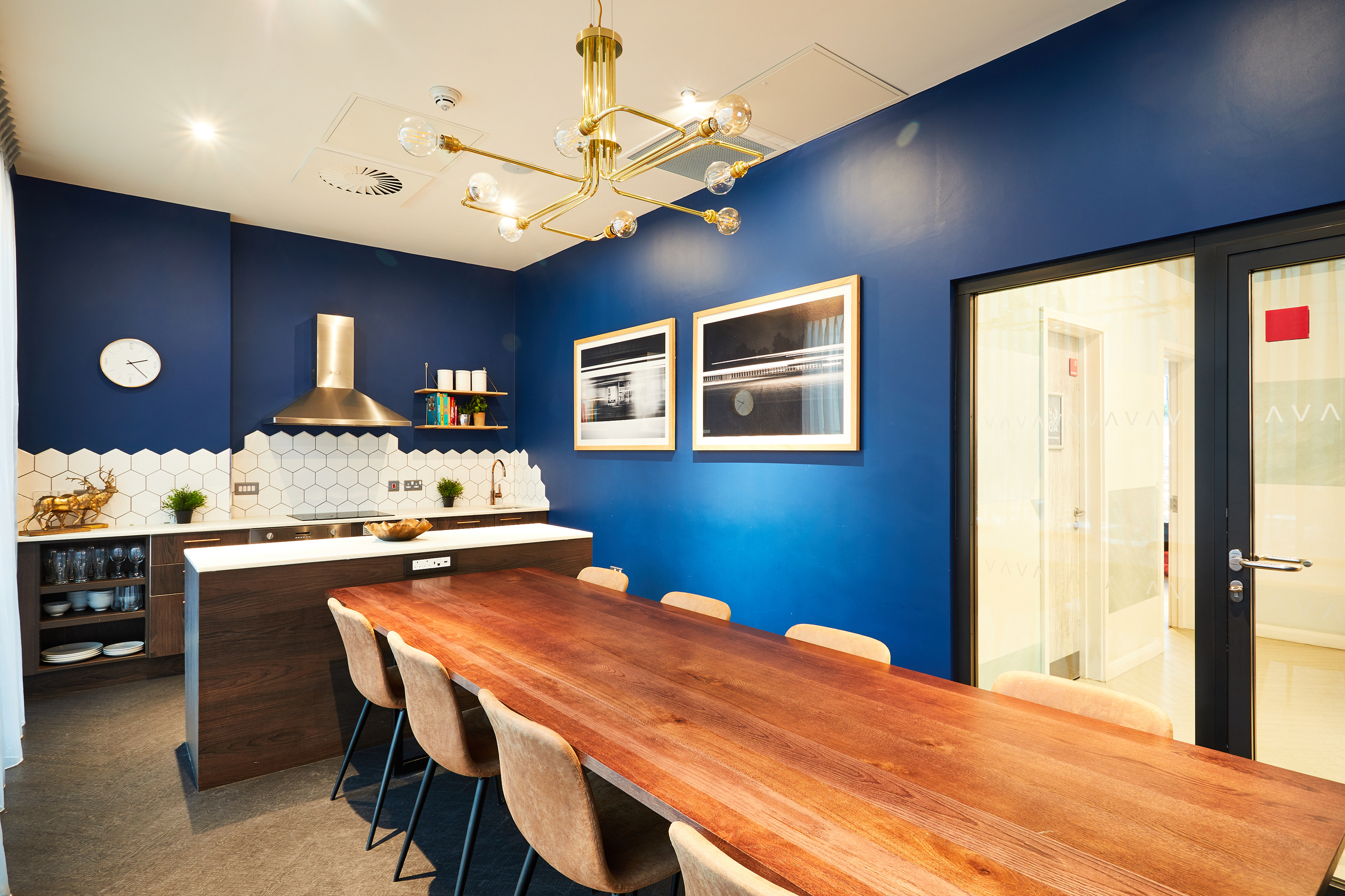 In the modern dining area of Strawberry Place, a long wooden table with eight chairs invites gatherings. Blue-painted walls are adorned with framed pictures. Nearby, the small kitchen boasts a hexagonal tile backsplash and shelves, while a gold light fixture elegantly illuminates the space.