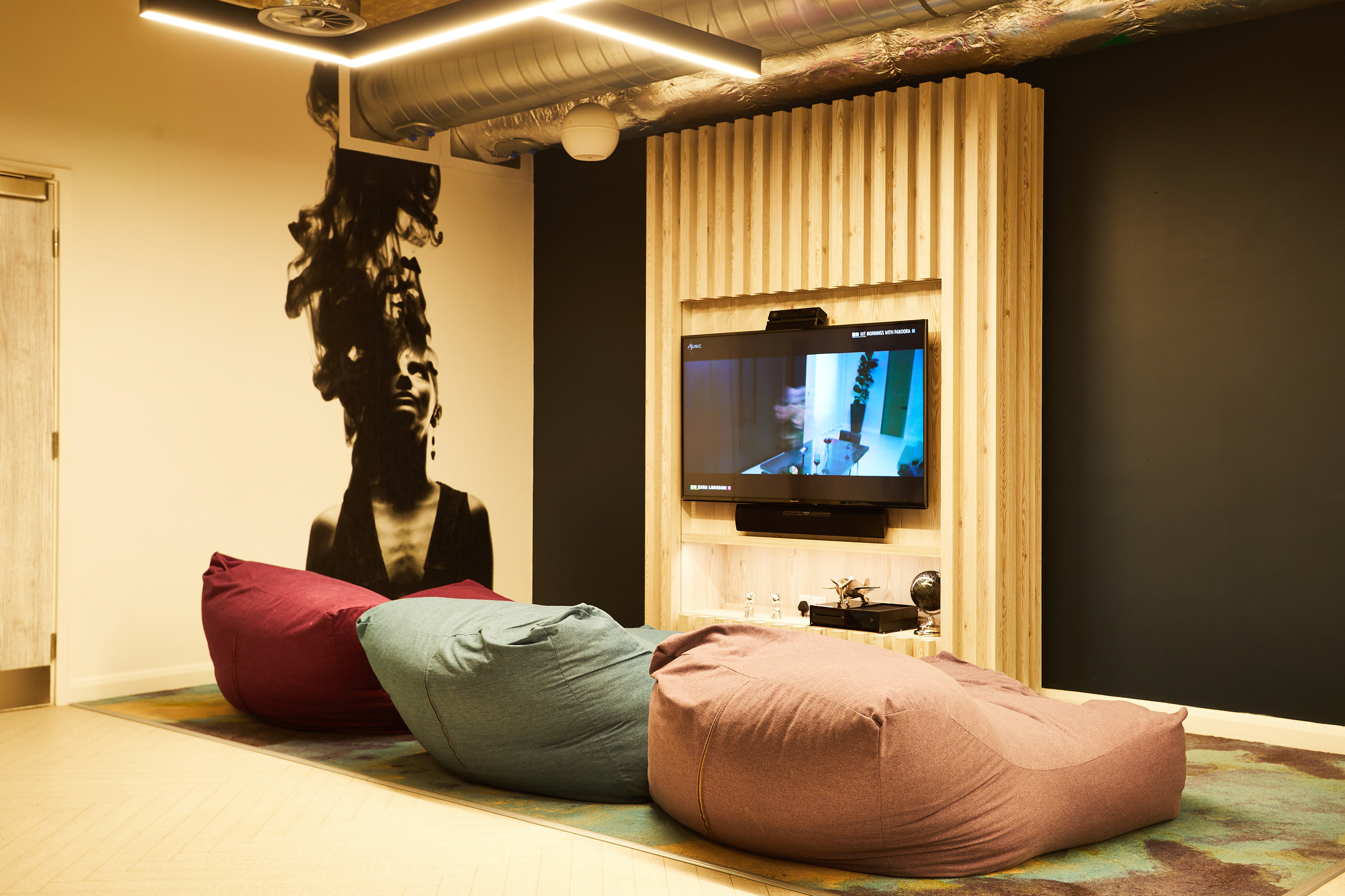 In the cozy room at Strawberry Place, a TV on a wooden accent wall displays a film. Three large bean bags in burgundy, teal, and purple rest on a rug. A mural of a person with abstract hair graces the wall, while modern lighting and an industrial ceiling complete the ambiance.