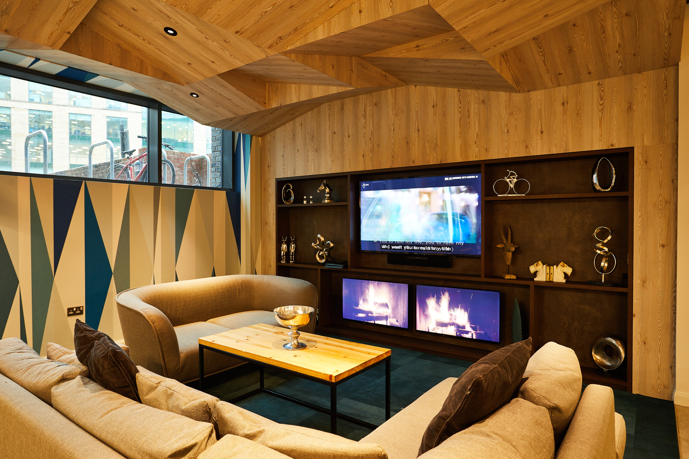 Cozy living room at Strawberry Place with wooden walls and ceiling, featuring a large TV above a dual-screen fireplace. Beige sofas surround a wooden coffee table. Decorative elements include shelves with art pieces and a large window offering views of bicycles outside.