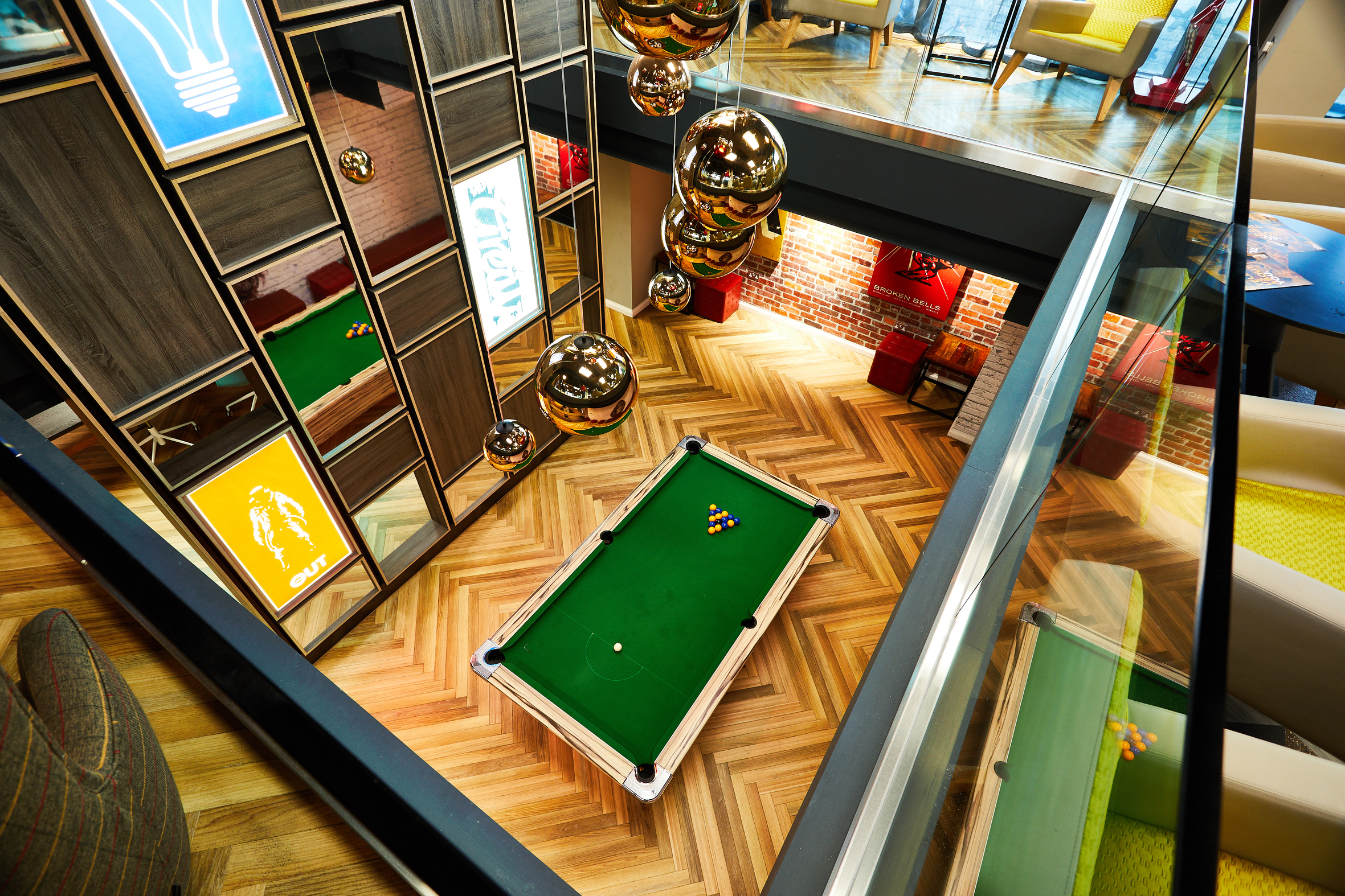 From a bird's-eye view, this stylish house showcases a modern room centered around a pool table. Wood flooring and glass railings complement colorful wall art and gold spherical hanging lights, while bright furniture adds vibrancy.