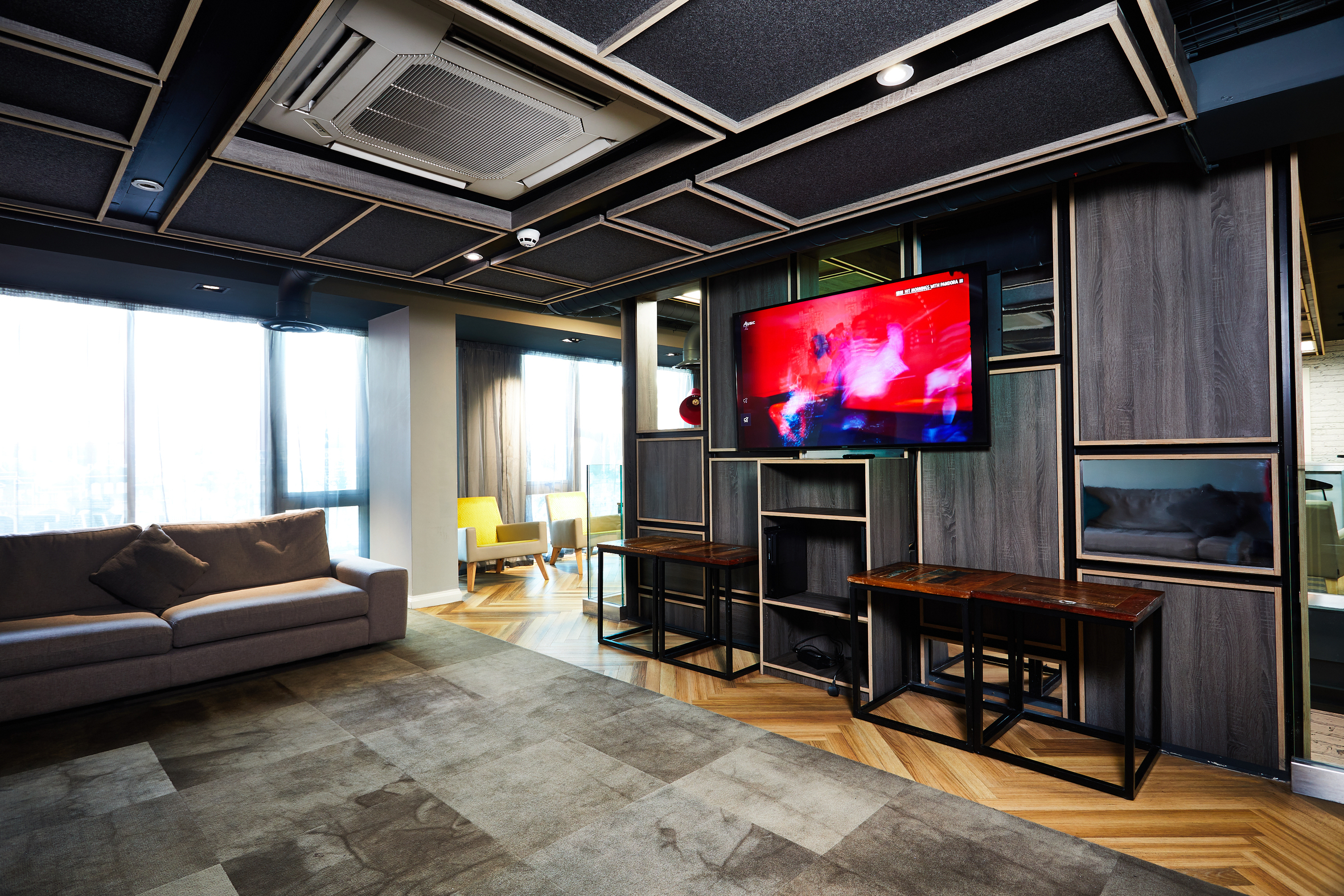 Modern living room in a stylish house with a large wall-mounted TV showing a colorful screen. The room features a sofa, two small wooden tables, and large windows allowing natural light. A telephone rests on one table beneath the decorative paneled ceiling, while the floor is a mix of wood and carpet.