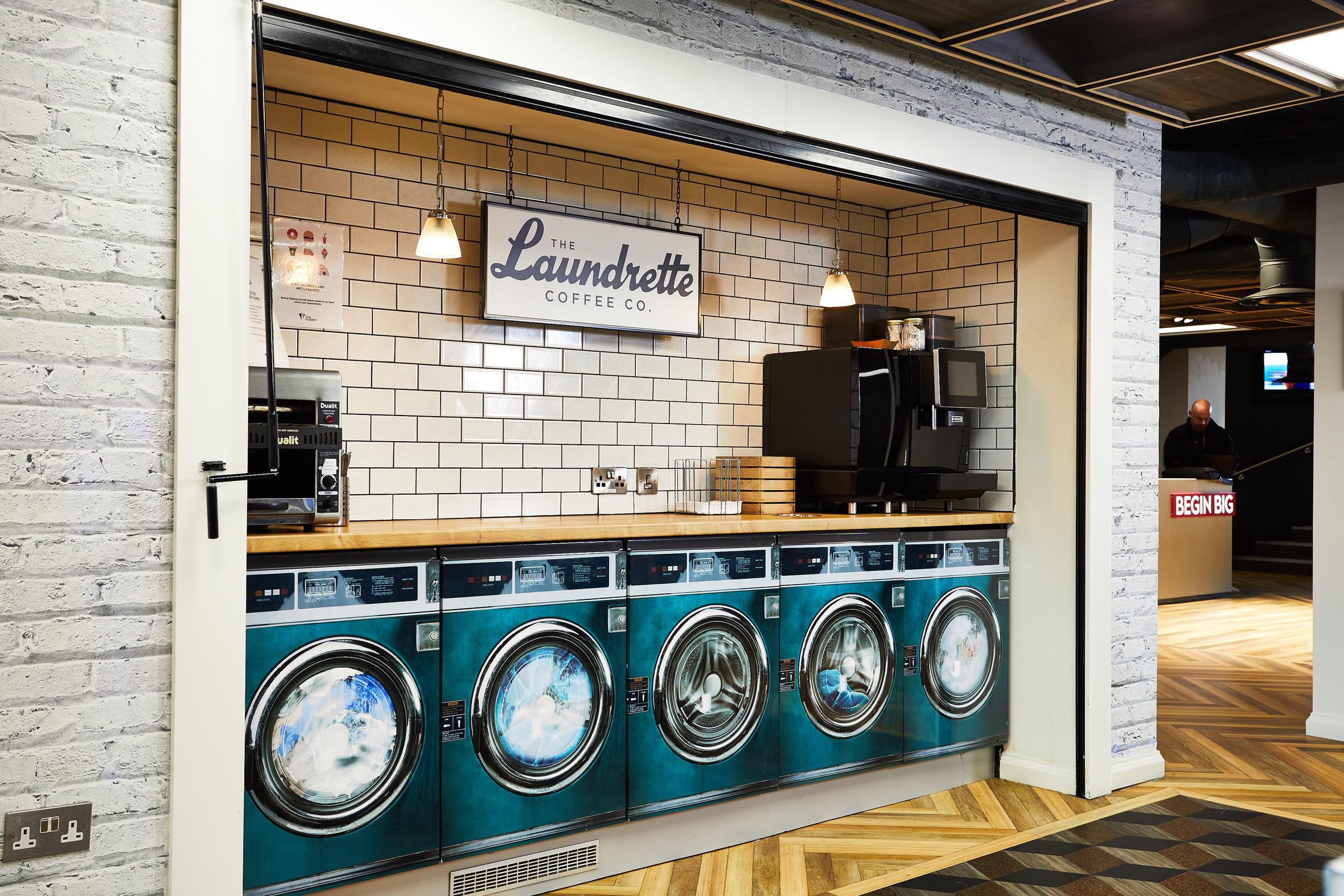 A cafe designed like a laundrette, with teal washing machines lined up below a wooden counter. A vintage telephone sits invitingly on the counter. A sign above reads "The Laundrette Coffee Co." The space features white tile walls and wooden herringbone flooring.