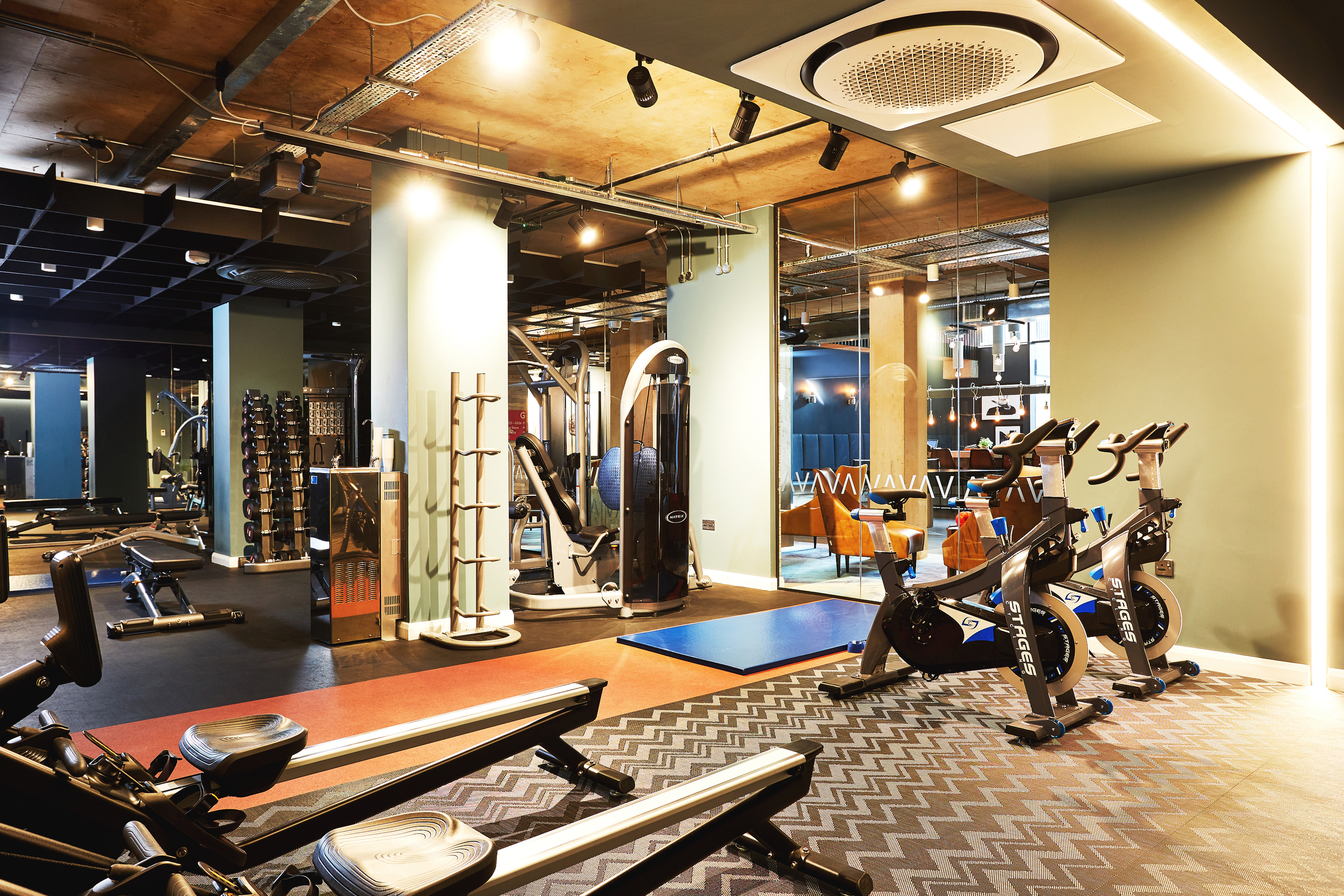 Modern gym interior at Portswood House in Southampton, featuring exercise bikes, weight racks, and various fitness equipment. The well-lit room with large mirrors and a wooden ceiling creates an inviting atmosphere for workouts. Perfect accommodation for fitness enthusiasts.