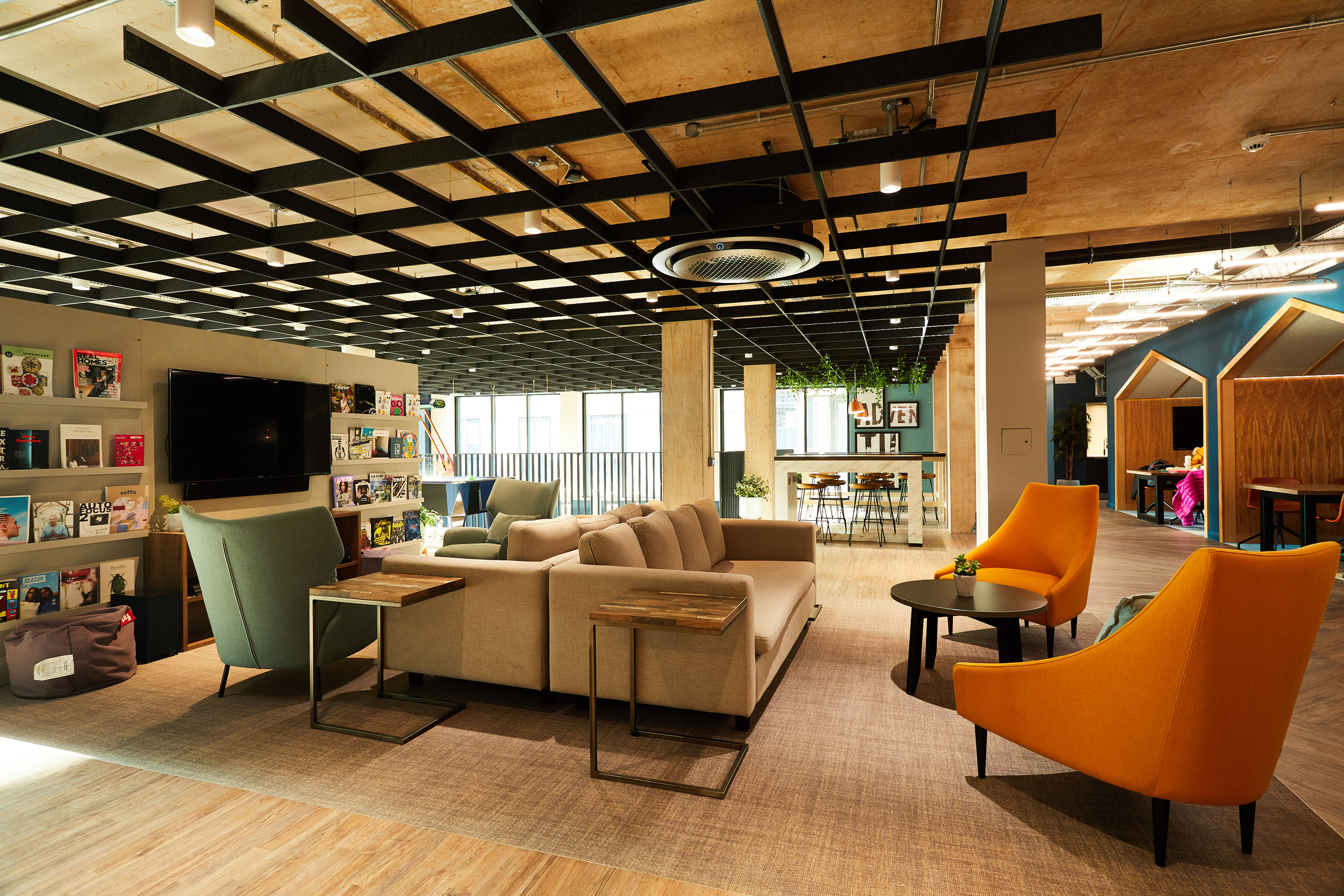 The modern office lounge at Portswood House boasts a cozy seating area with a beige sectional sofa, green and orange chairs, and small tables. Bookshelves and a TV adorn the background, as natural light streams through the windows beneath a black grid ceiling.