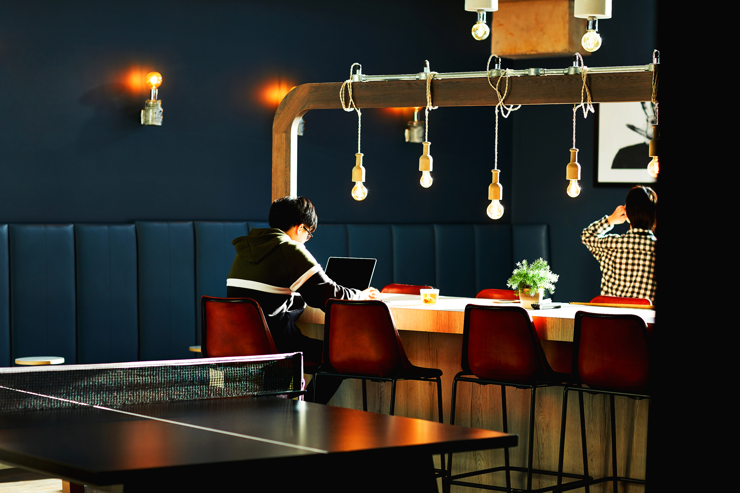Two people sit at a long table with hanging lights in a modern, dark-walled room in Portswood House. One works on a laptop while the other adjusts their hair, capturing the essence of student accommodation life. A ping pong table is visible nearby, and a small potted plant decorates the scene.