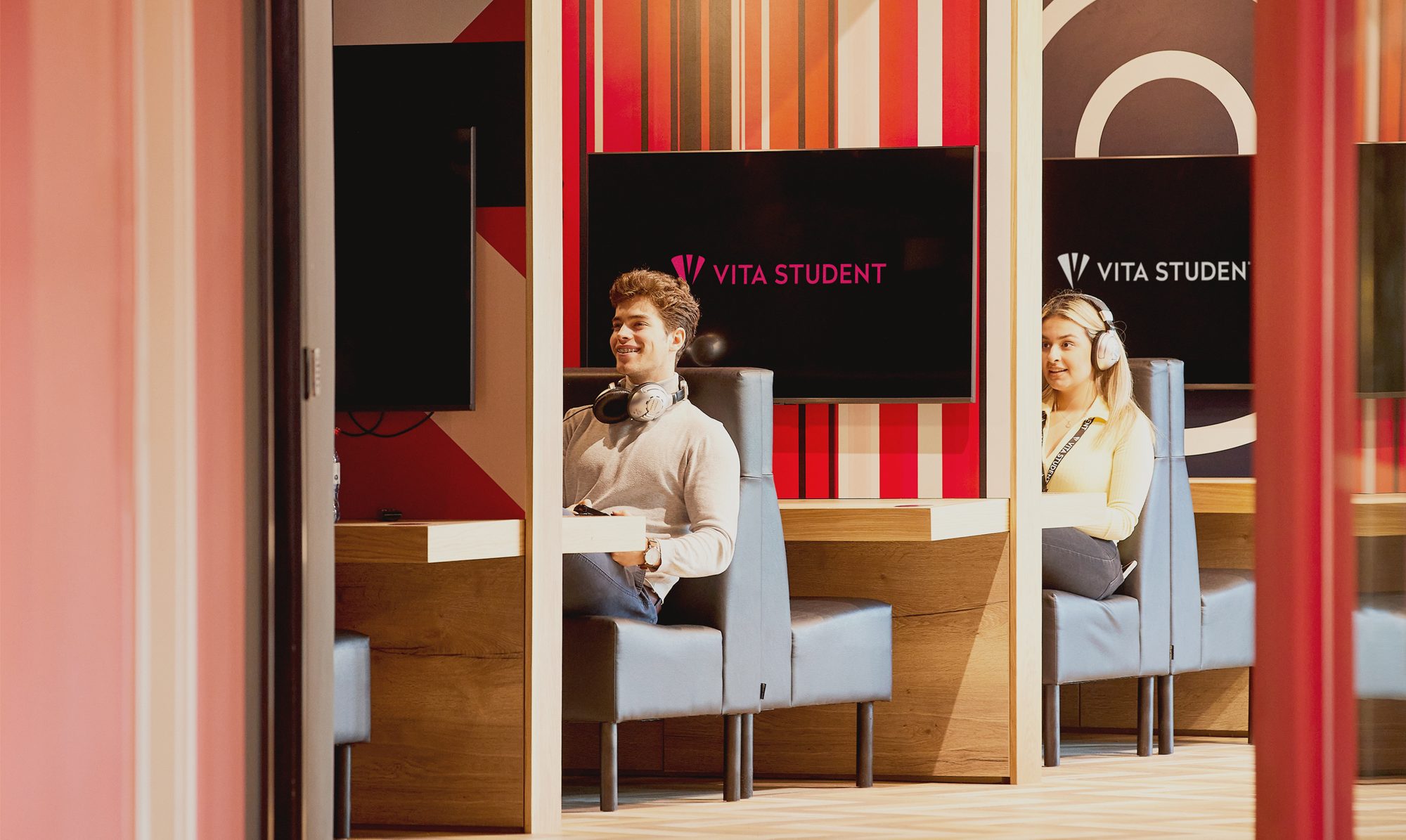 Two people sit separately in brightly colored study booths with headsets on. Each booth has a screen displaying "Vita Student." The background features a pattern of red and orange stripes.