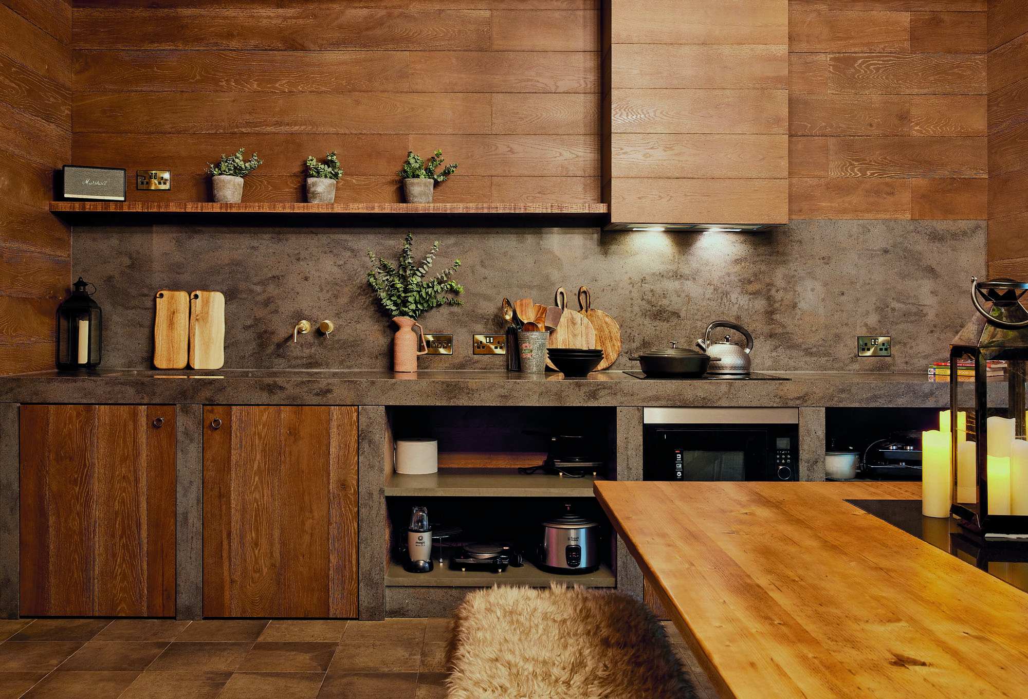 A rustic kitchen with wooden cabinets and a concrete countertop, reminiscent of the charming styles found in Lewisham Exchange. Potted plants, cutting boards, and cooking utensils grace the counter. A metallic kettle enhances the charm, while a furry chair invites comfort near a wooden table.