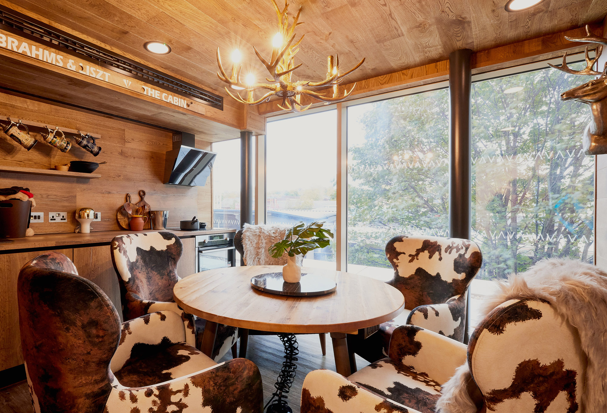 The rustic dining room at Lewisham Exchange boasts a wooden ceiling and walls, with a round wooden table surrounded by cowhide-patterned chairs. Large windows offer views of trees outside, while an antler chandelier hangs above a small plant centerpiece.