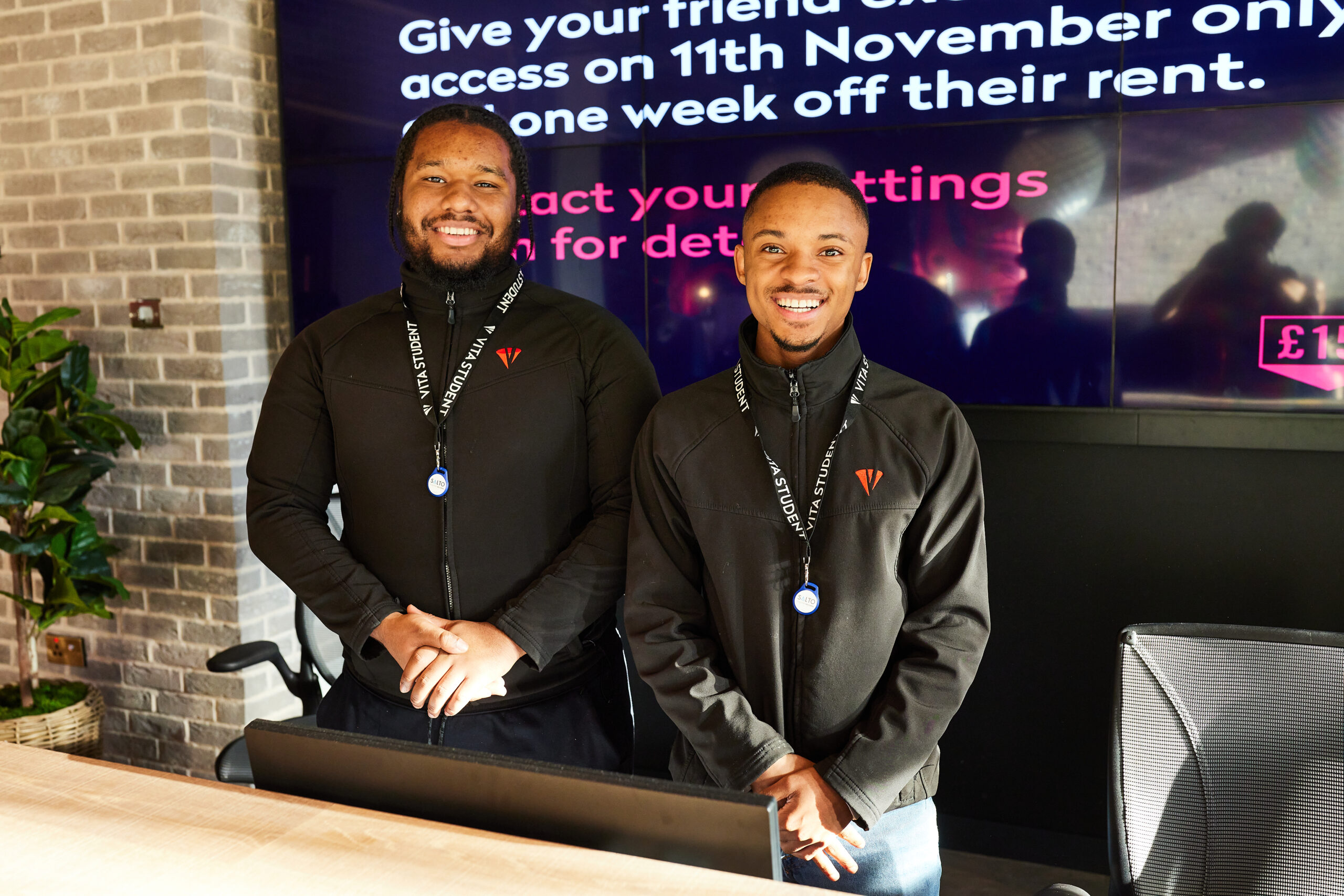 Two smiling men stand behind a desk, both wearing black jackets and lanyards. A large screen with text adds life to the background, while part of a brick wall is visible on the left. The setting appears to be an indoor office or reception area.