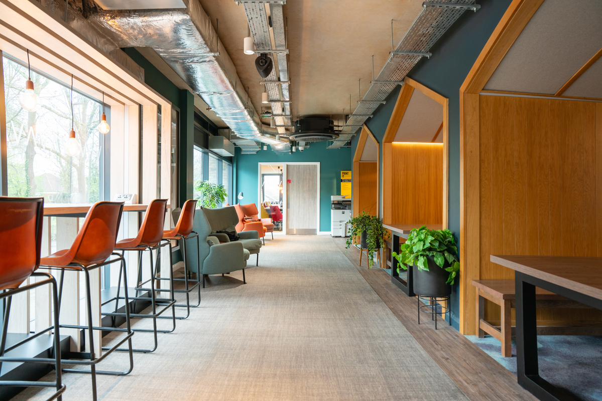 Portswood House offers a modern coworking space with high and low seating areas. The left side features tall bar stools and a counter by the window, while the right side boasts cozy chairs and tables with plants. Exposed ceilings and wooden accents add an industrial feel to this dynamic workspace.