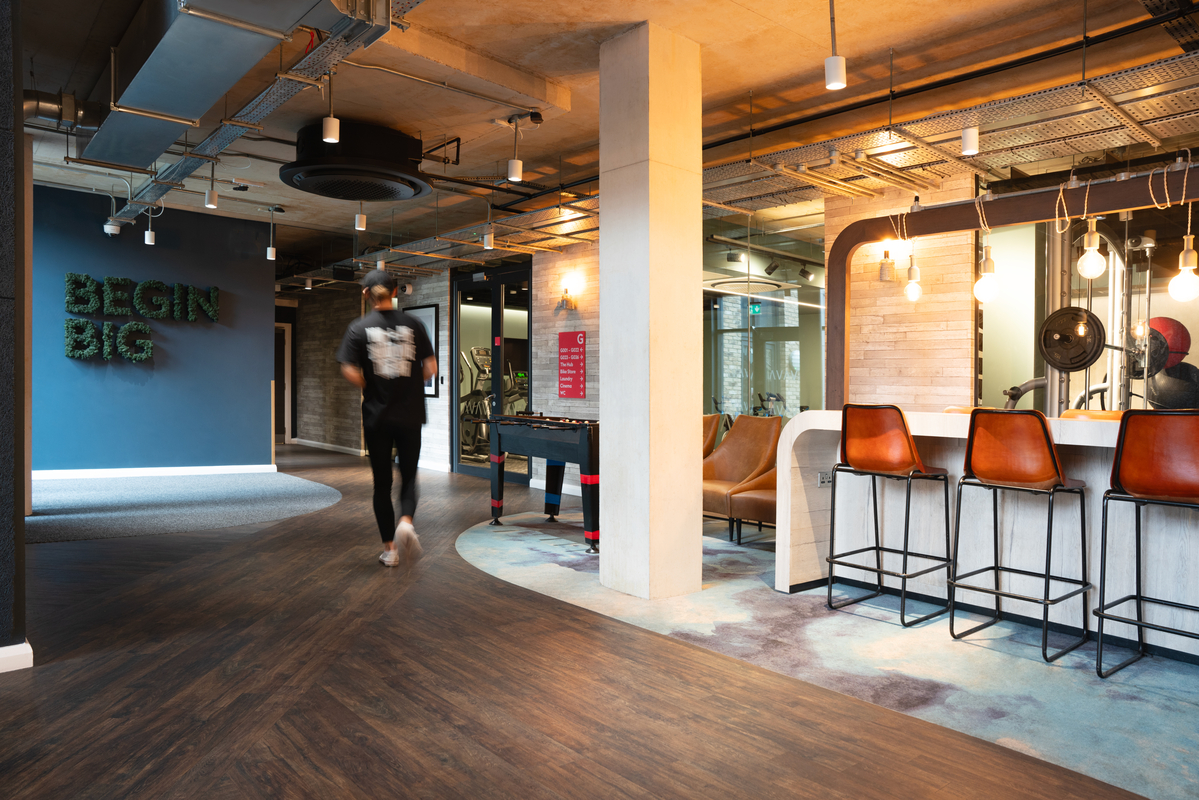 Modern lounge area at Portswood House with a bar featuring four high chairs, a foosball table, and decorative lighting. A person walks across the wooden floor. The blue wall displays the phrase "BEGIN BIG" in greenery. Exposed ceiling with visible ducts adds an industrial touch.