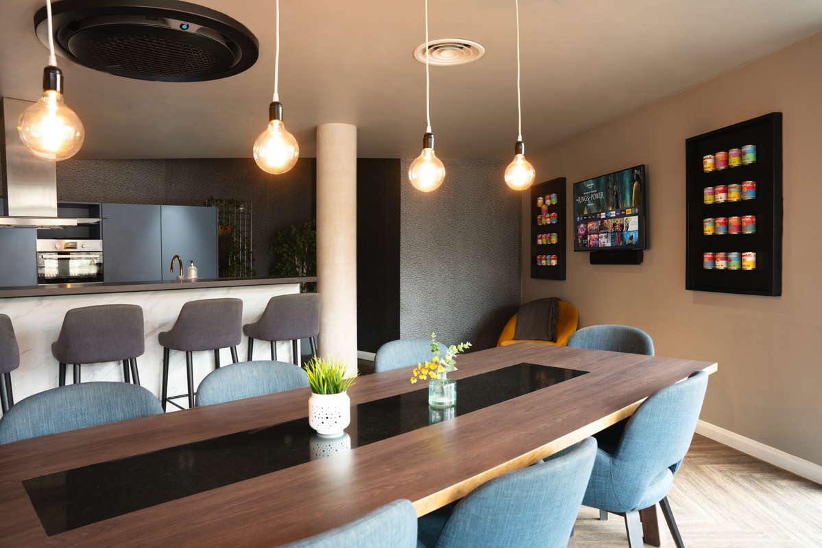 Modern dining area within Portswood House features a wooden table surrounded by blue chairs. Three pendant lights hang above, illuminating the student housing space. In the background, a kitchen island with barstools, a wall-mounted TV, and colorful decor jars add charm. A small plant adorns the table.