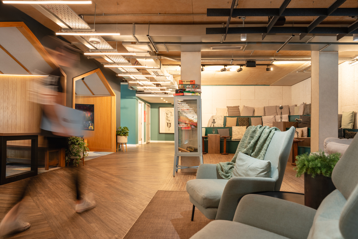 The modern office space at Portswood House features armchairs in the foreground, books and games on shelves, and cushioned seating along the back wall. A person briskly walks by, creating a motion blur. The decor is warm and inviting with wooden and green accents.