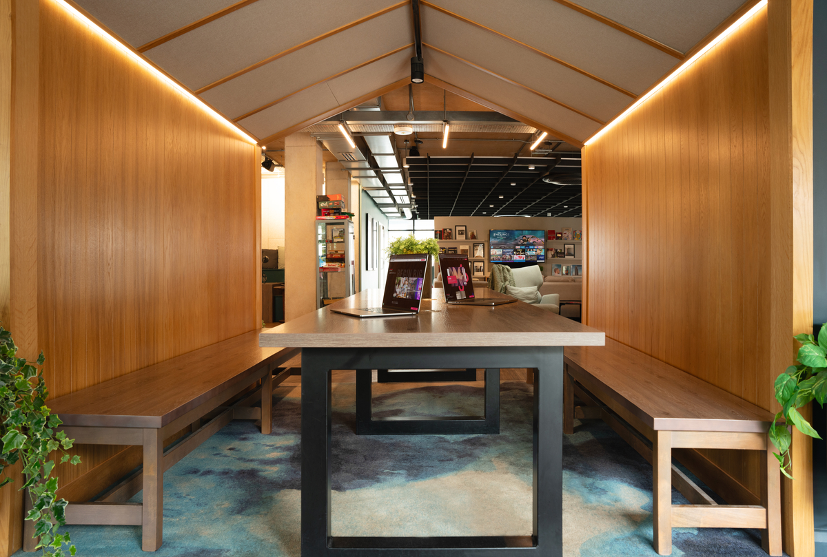 A modern office space in Portswood features a long wooden table with benches, enclosed within a house-like structure with a pitched roof. Wooden walls and ceiling complement the hanging plants, while a patterned rug adds warmth to the floor.