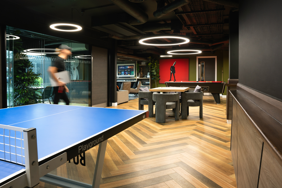 A modern recreational room in student accommodation showcases a blue ping pong table in the foreground. In the background, there's a pool table and lounge area with a television. A person walks by, while circular ceiling lights illuminate the herringbone wood flooring.