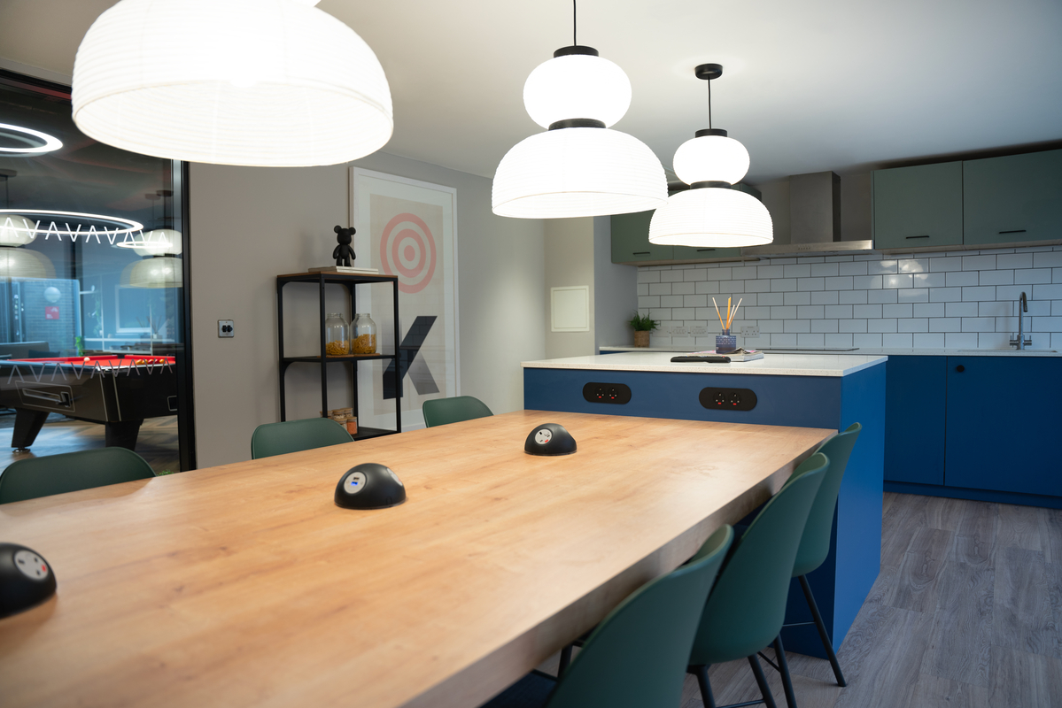 The modern student accommodation kitchen boasts a wooden dining table, green chairs, and three large pendant lights. Blue cabinets and a white subway tile backsplash complement the room, while a pool table is visible through the glass door for leisure activities.