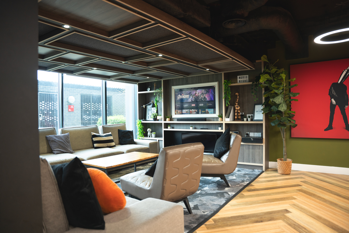 This modern lounge, perfect for student accommodation, features a sectional sofa, armchairs, and a wooden coffee table on a patterned rug. A TV adorns the wall alongside bookshelves and plants, while striking red artwork adds flair. Large windows flood the space with natural light.