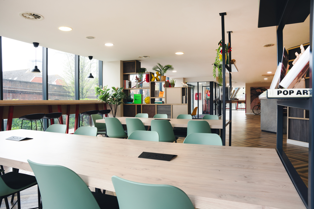 Modern, bright workspace reminiscent of student accommodation, featuring long tables with light wood surfaces and pastel green chairs. Large windows on one side let in natural light. Shelves contain books and plants, complemented by ceiling lighting and wood-patterned flooring.