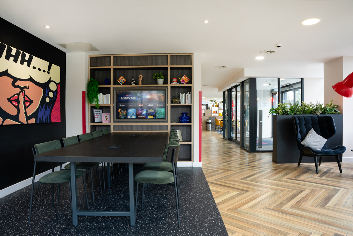 In this student accommodation, a modern office lounge features a long table, green chairs, and artwork on the wall. Bookshelves display various items, while a cozy chair with cushions sits in the corner. The herringbone-patterned floor leads seamlessly to glass-partitioned offices.