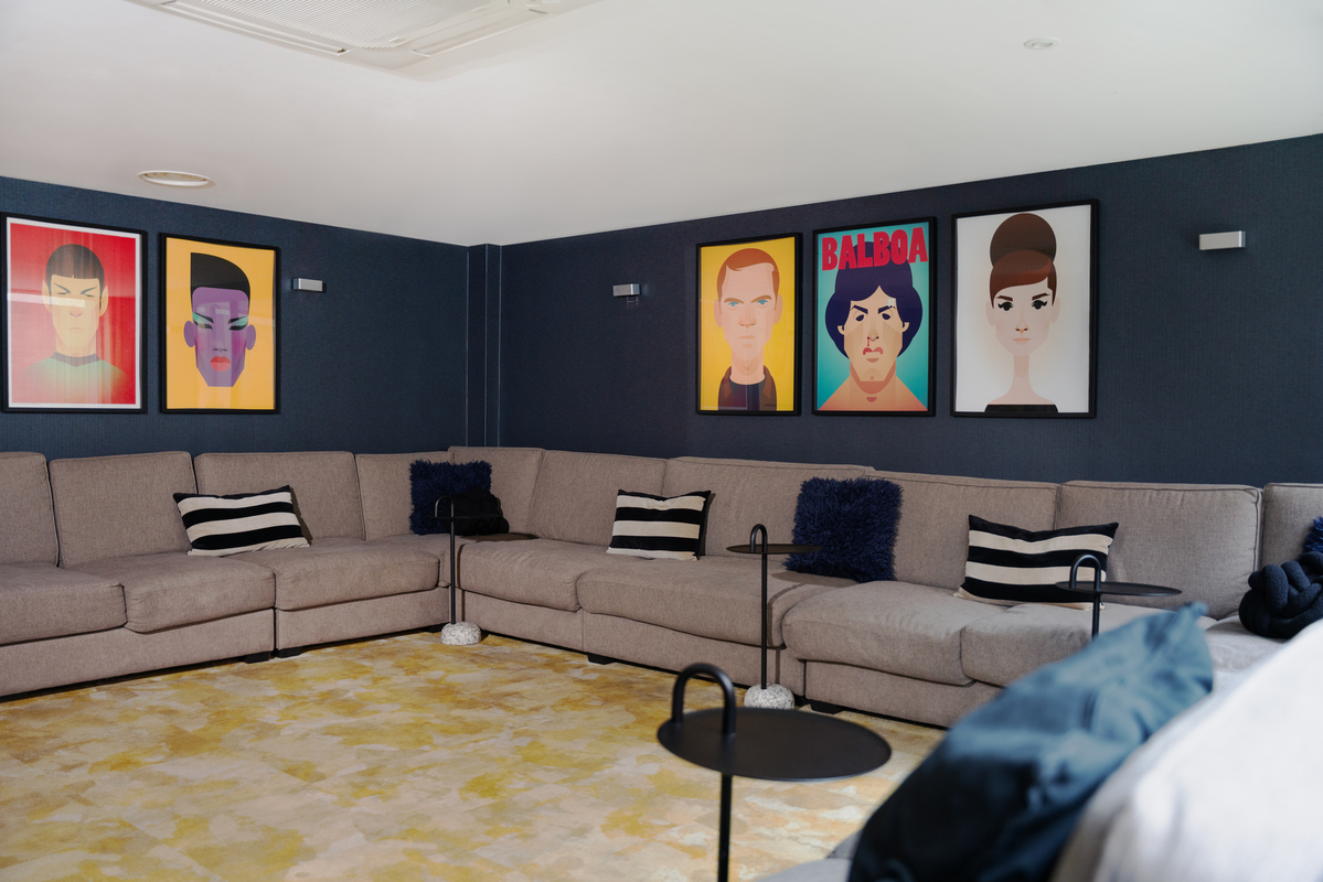 A modern lounge in the student accommodation features a large, U-shaped beige sofa with striped cushions. Four colorful, stylized portraits adorn the dark walls, while a textured yellow rug and round black tables complete this cozy setting.