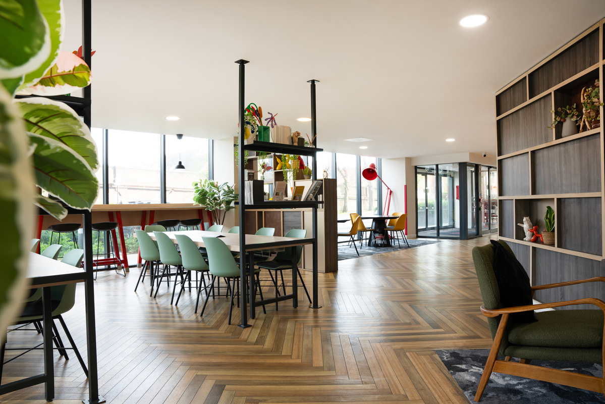 Modern office space with wooden herringbone flooring, featuring tables and assorted chairs, large windows, and bright red and green accents. Shelving displays decor items and plants, while a lounge area with an armchair adds a cozy, student accommodation-inspired touch.
