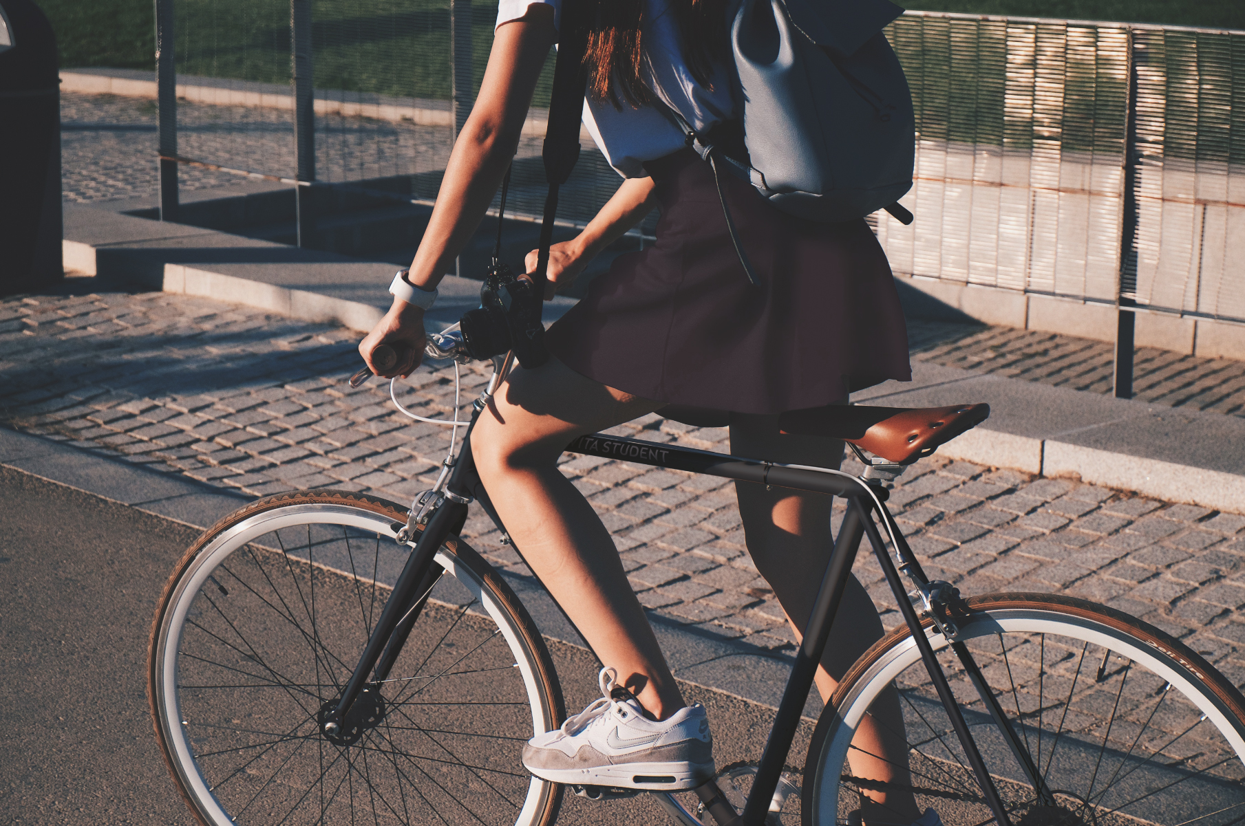 An international student rides a bicycle on a paved path, wearing a dark skirt and white sneakers, with a backpack slung over their shoulder. In the background, a grassy area and metal fence offer a sense of adventure in this new environment.