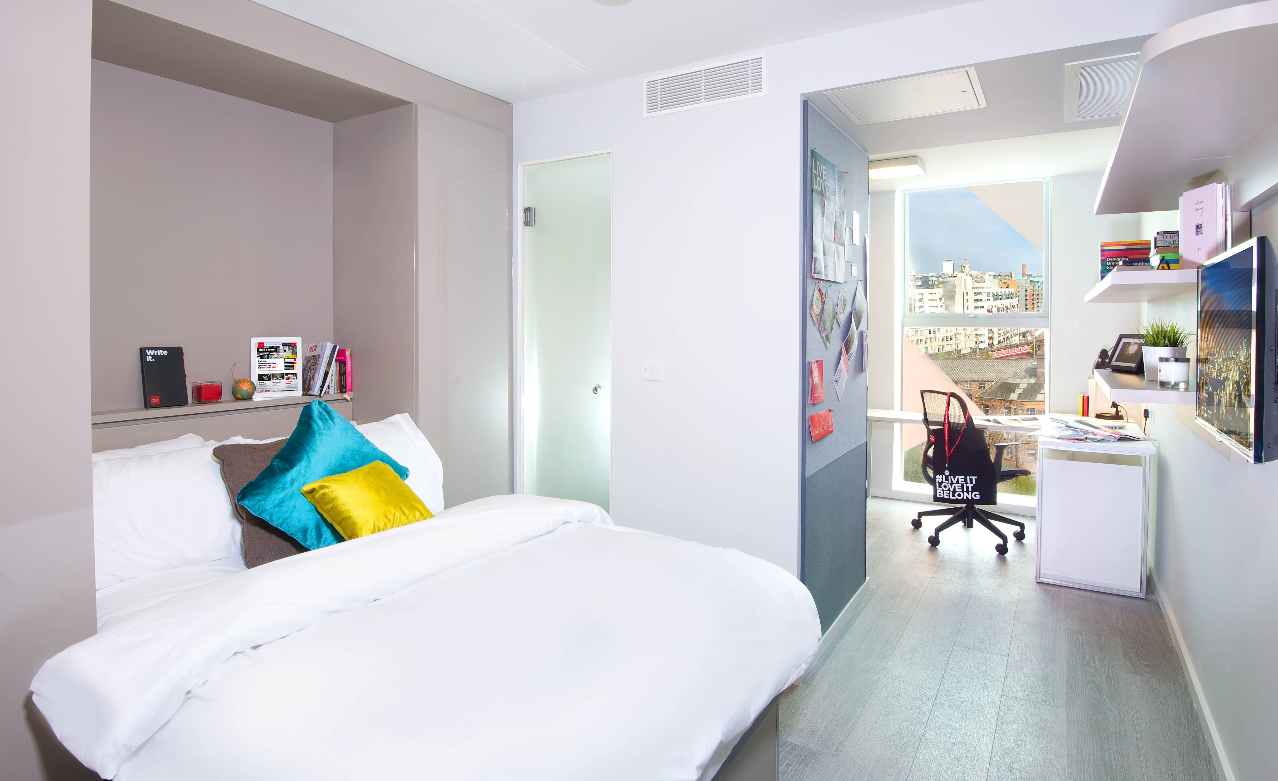 A bright, modern bedroom on First Street features a neatly made bed with colorful pillows on the left. A desk with a chair sits near the window, offering a view of city buildings. Shelves and a wall-mounted TV are on the right. The room boasts a sleek, minimal design.