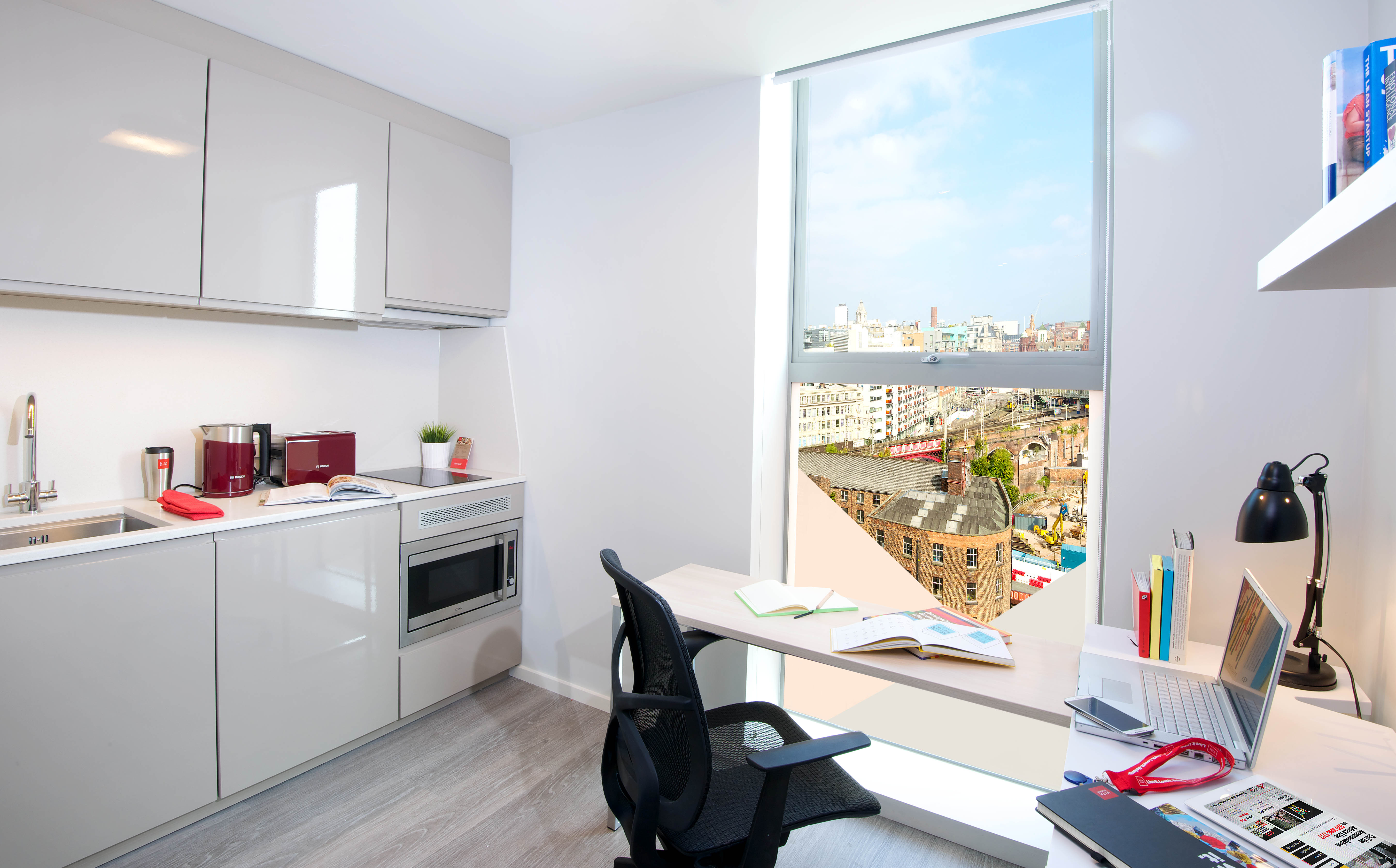 This modern apartment on First Street boasts a kitchenette with a microwave, sink, and cabinets. A desk equipped with a laptop, notebooks, and lamp complements the spacious room. Enjoy the cityscape through a large window that illuminates the light wood flooring beautifully.