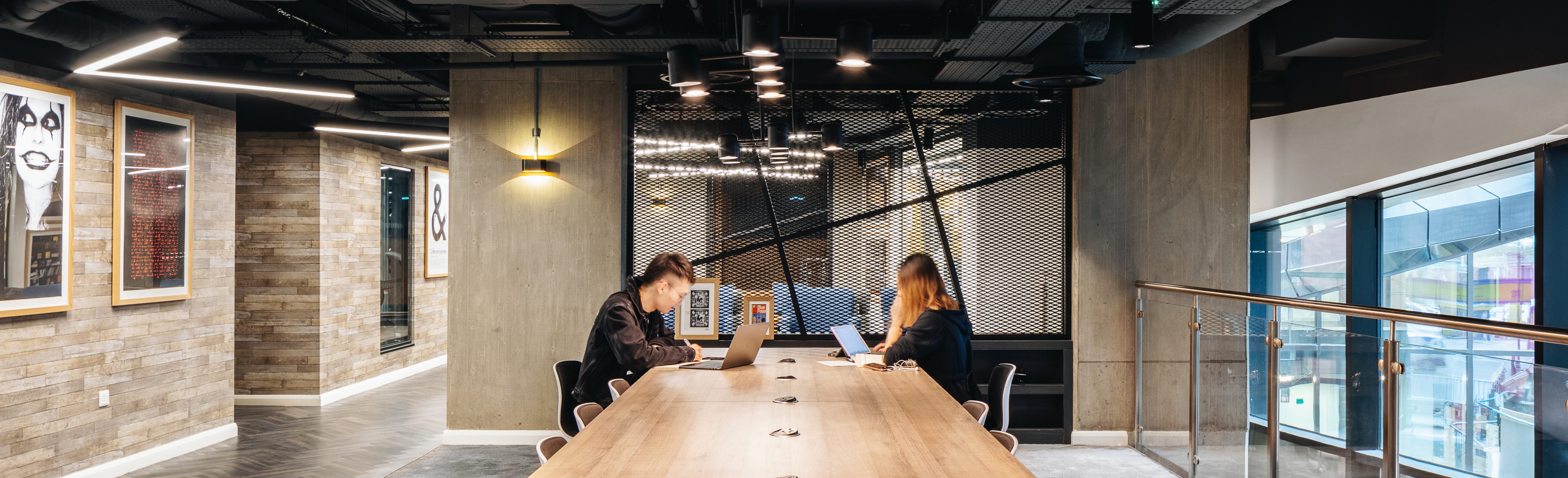 Two people work on laptops at a long wooden table in a modern office space, mindful of the company's privacy policy. The room features exposed ceilings, brick walls, framed artwork, and large windows. Hanging lights illuminate the scene, while a staircase is visible in the background.