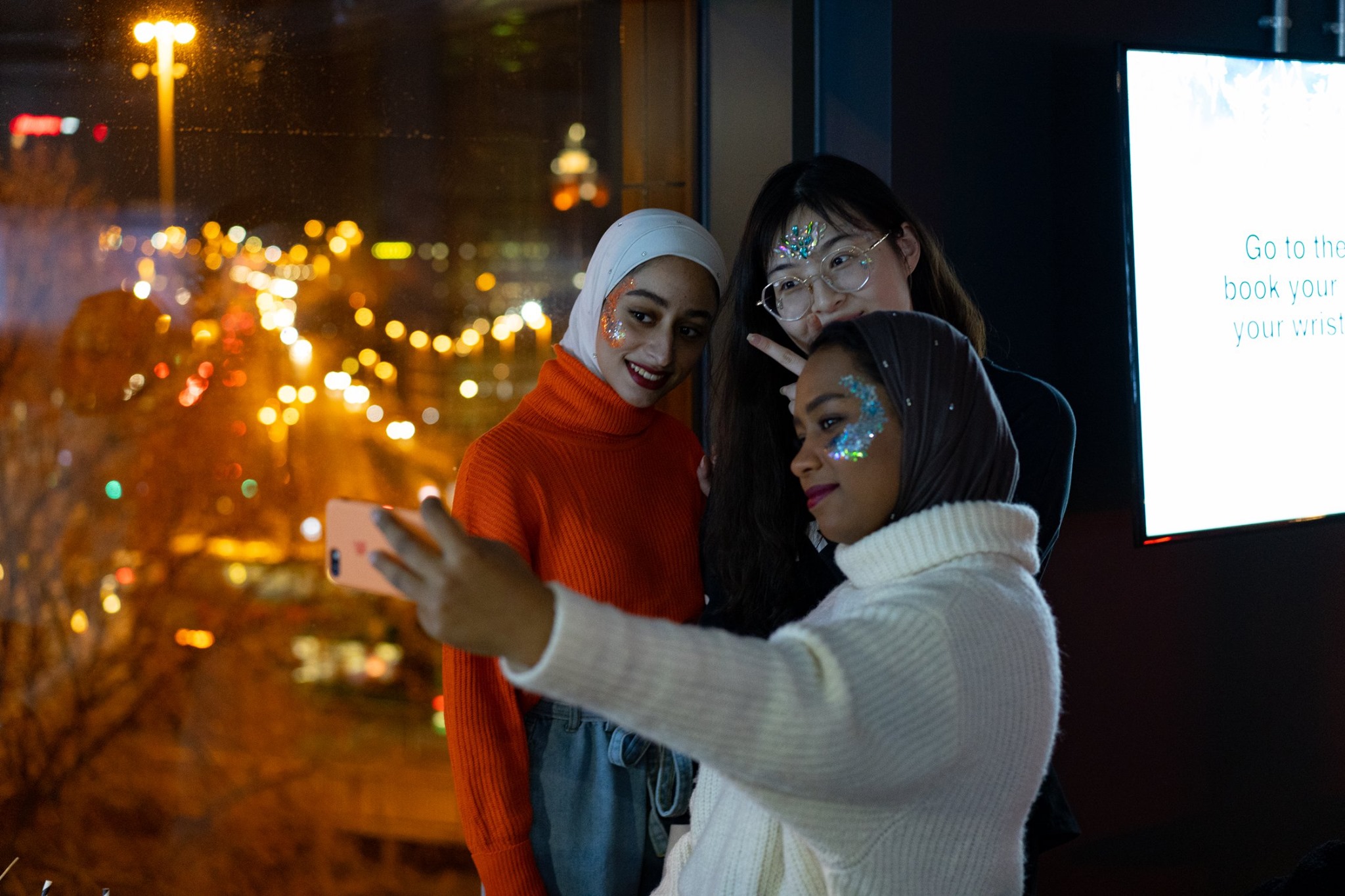 Three international students pose for a selfie near a window with a city nightscape in the background. They are smiling, with glitter on their faces. One wears a white hijab and red sweater, another has glasses, and the third is in a white turtleneck.