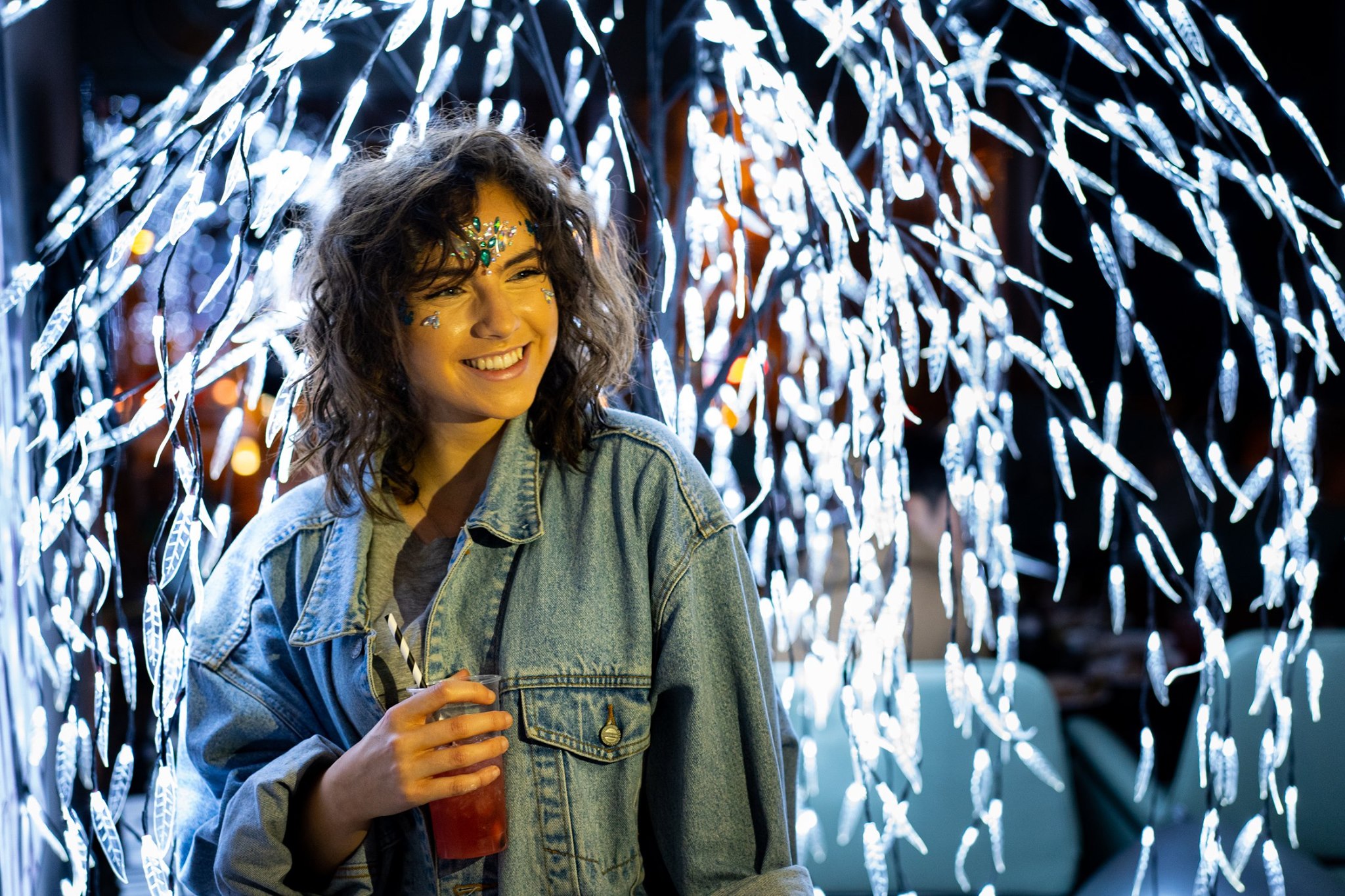 A woman with curly hair and glitter makeup smiles while holding a red drink, exuding a distinctly English charm. She wears a denim jacket and stands in front of a display of bright, cascading lights reminiscent of festive UK streets.