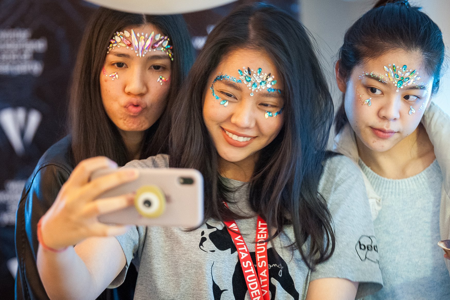 Three people taking a selfie, each with colorful, jeweled face decorations. They are smiling and posing together—one making a pouty face. The setting gives the vibe of an English pub with dim lighting, suggesting they're having a night out in the UK.