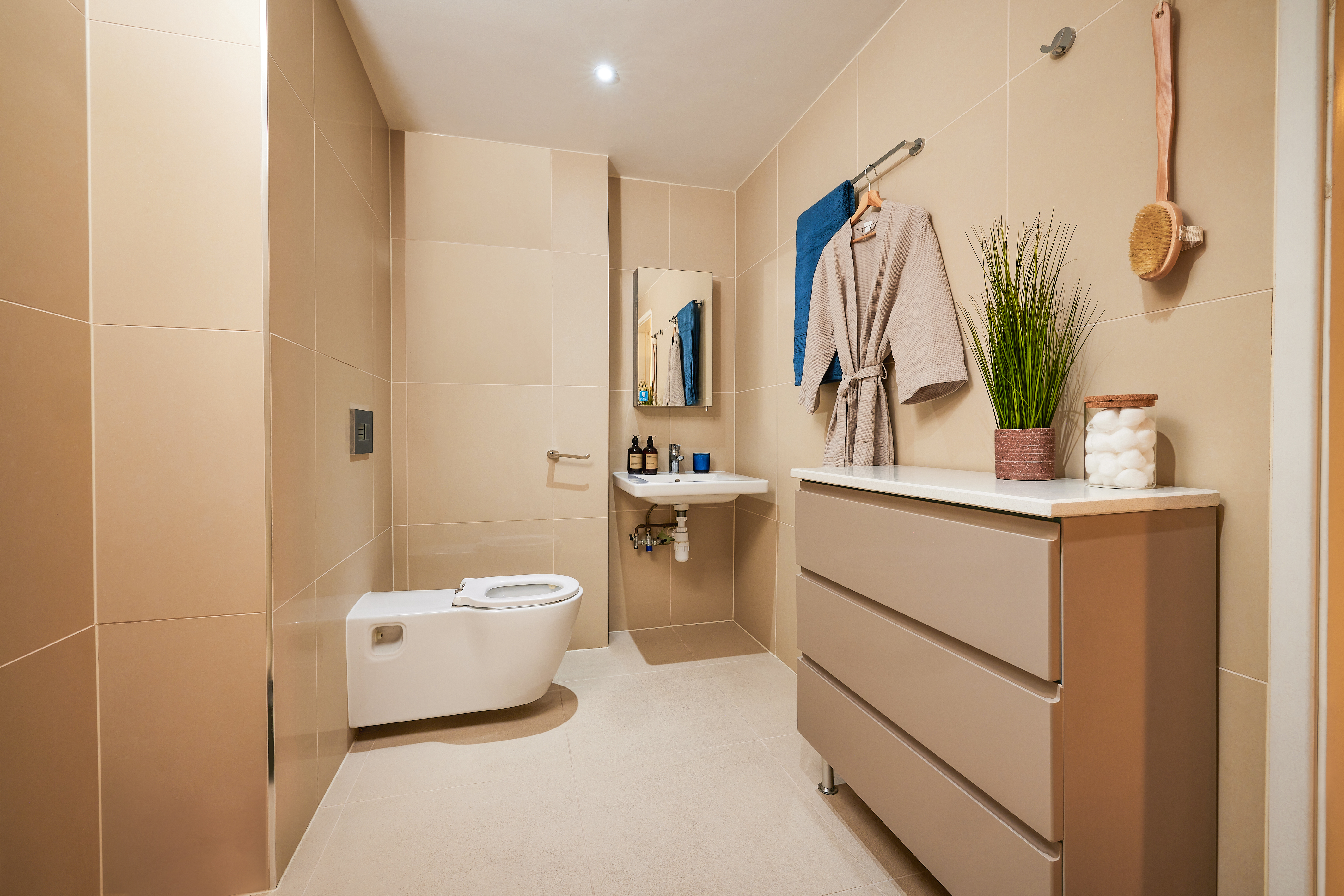 Modern bathroom with beige tiles, a wall-mounted toilet, and a small sink. A grey robe hangs on the wall beside a mirror. A chest of drawers holds a potted plant and jar of cotton balls. A brush hangs beside a blue towel.