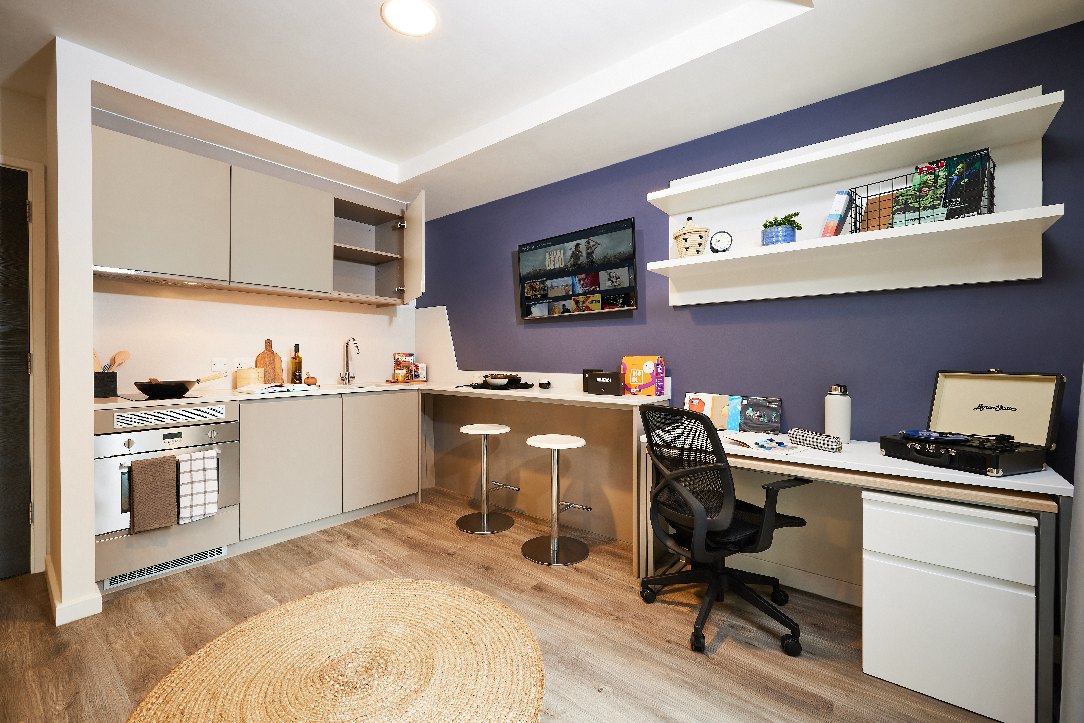 A modern studio interior with a small kitchen on the left, featuring cabinets, a stove, and a sink. On the right, there's a desk with a laptop and shelves above. A wall-mounted TV is in the center. The floor is wooden with a round rug.