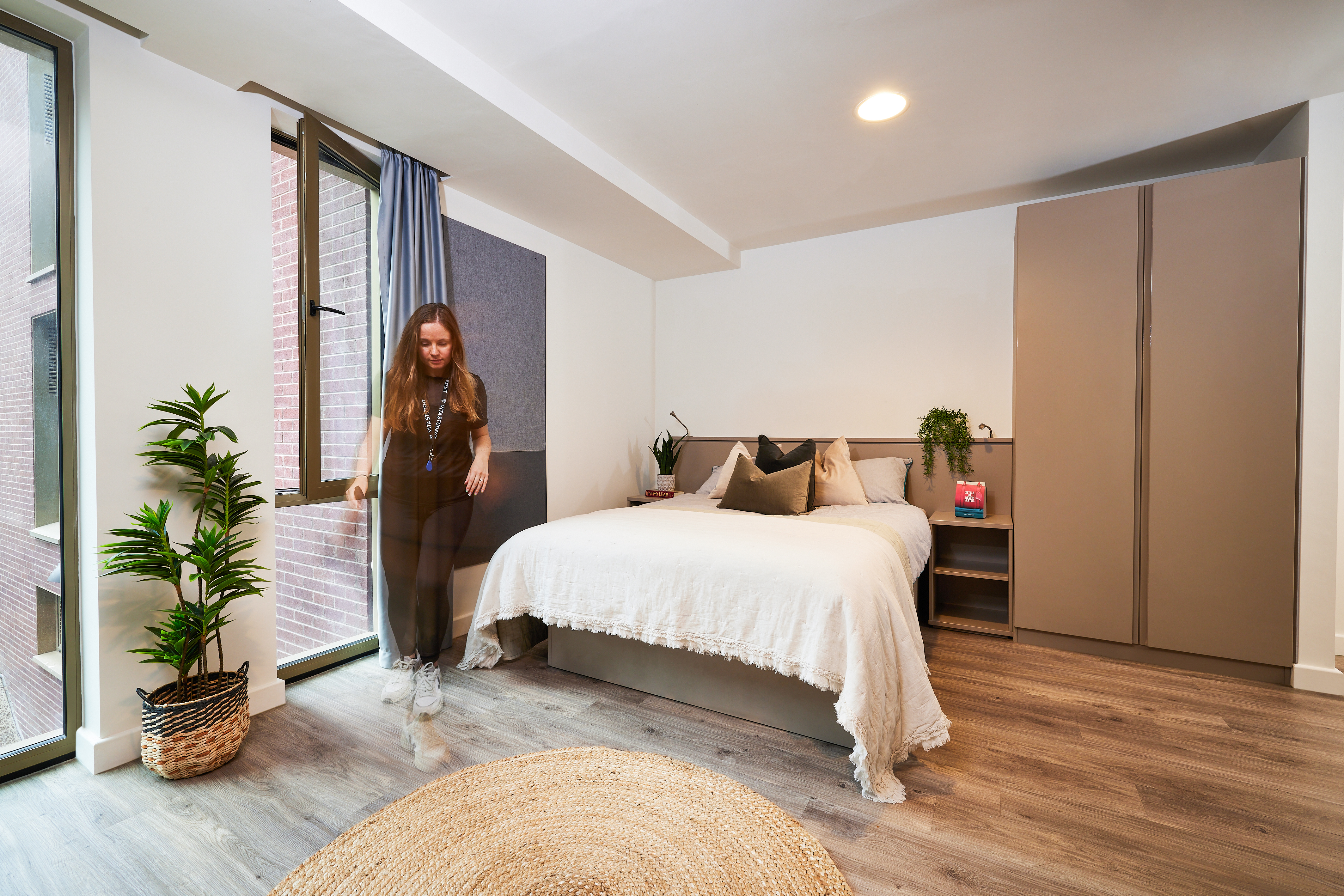 A bedroom with a bed, nightstand, and wardrobe. The bed has pillows and a white blanket. A person walks near large windows with blue curtains. A potted plant and woven rug adorn the wooden floor. Brick wall visible through the windows.