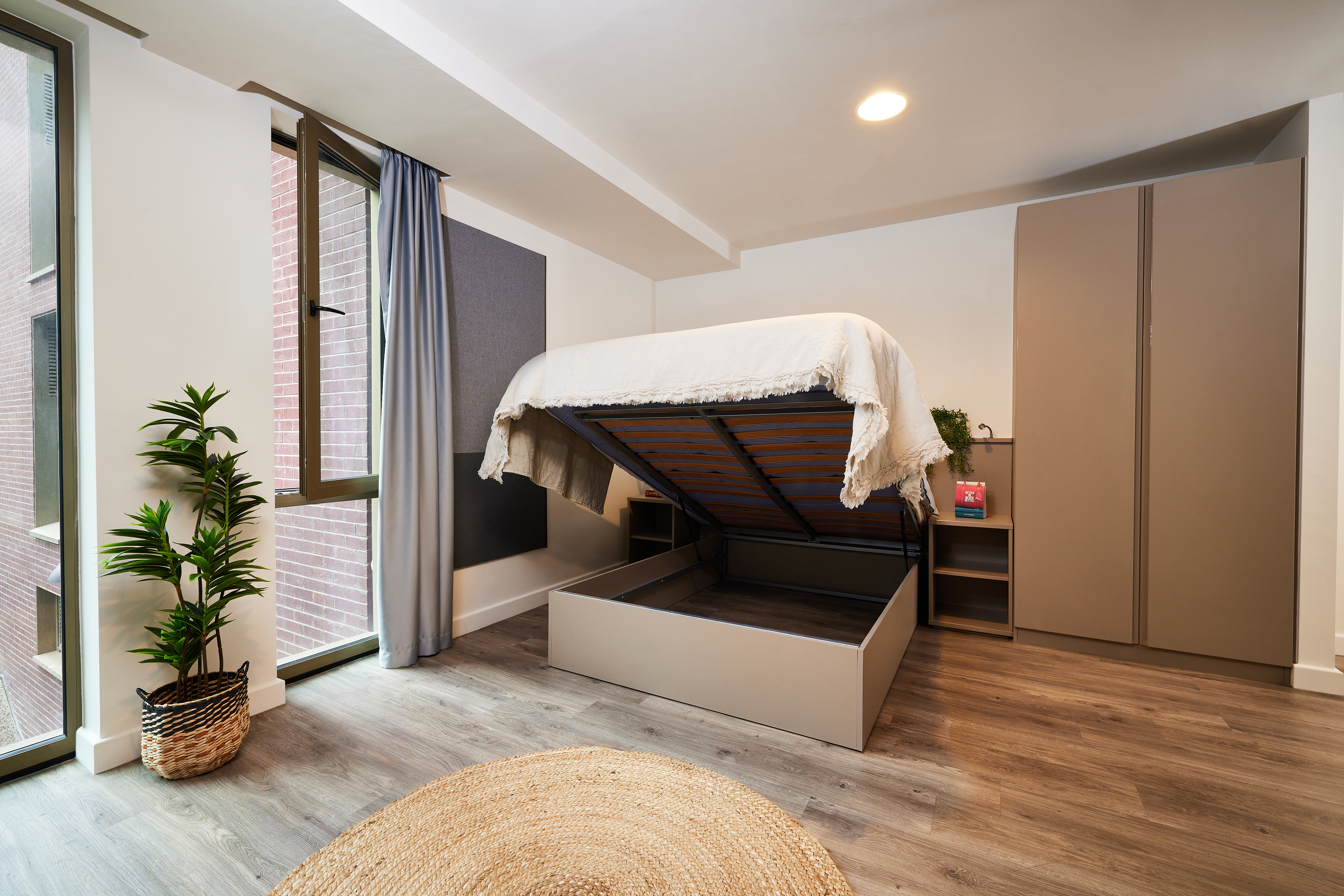 Modern bedroom with wooden flooring featuring a bed with storage underneath, shown open. A wardrobe is in the corner, and a small nightstand with a plant and book is beside it. Light streams in through a window, and a round rug is on the floor.