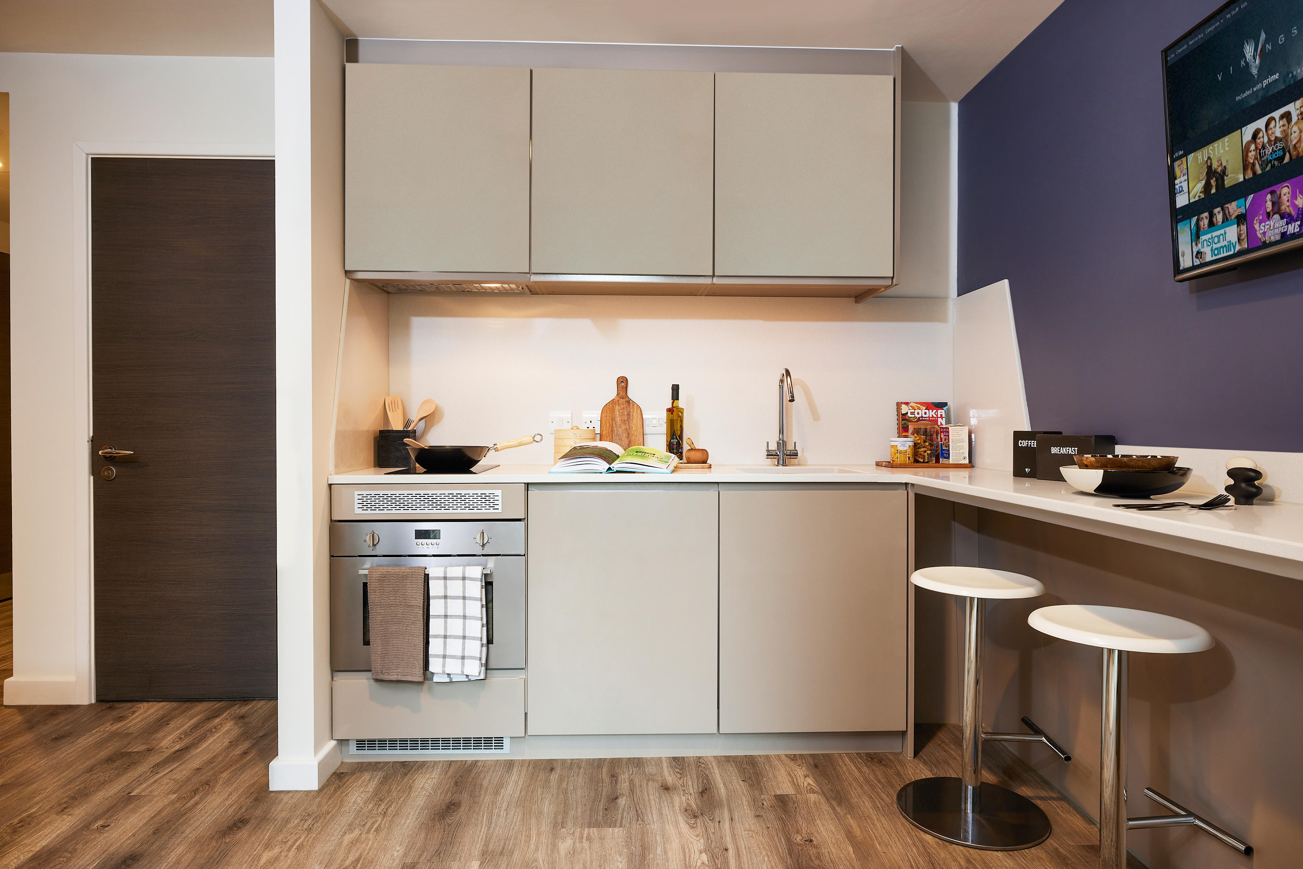 A modern kitchen with beige cabinets, an oven, and a stainless steel sink. A cookbook and utensils sit on the counter. Two bar stools are next to a white counter, with a TV mounted on the purple wall. Laminate wood flooring completes the space.