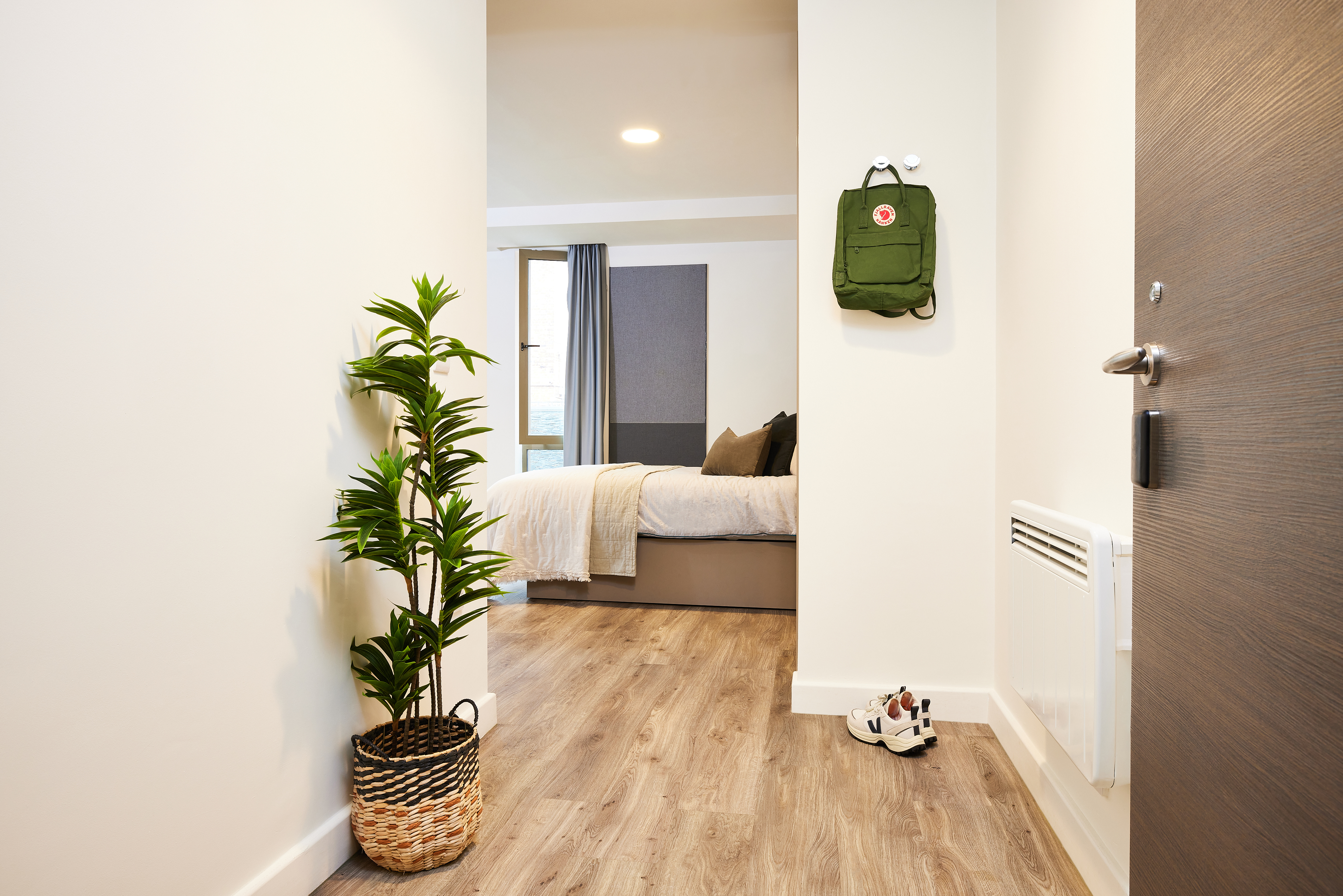 View of a minimalist bedroom from a hallway, featuring a neatly made bed with neutral linens. A potted plant sits on the wooden floor in the hallway, which has a green backpack hanging on the wall and a pair of shoes near the entrance.