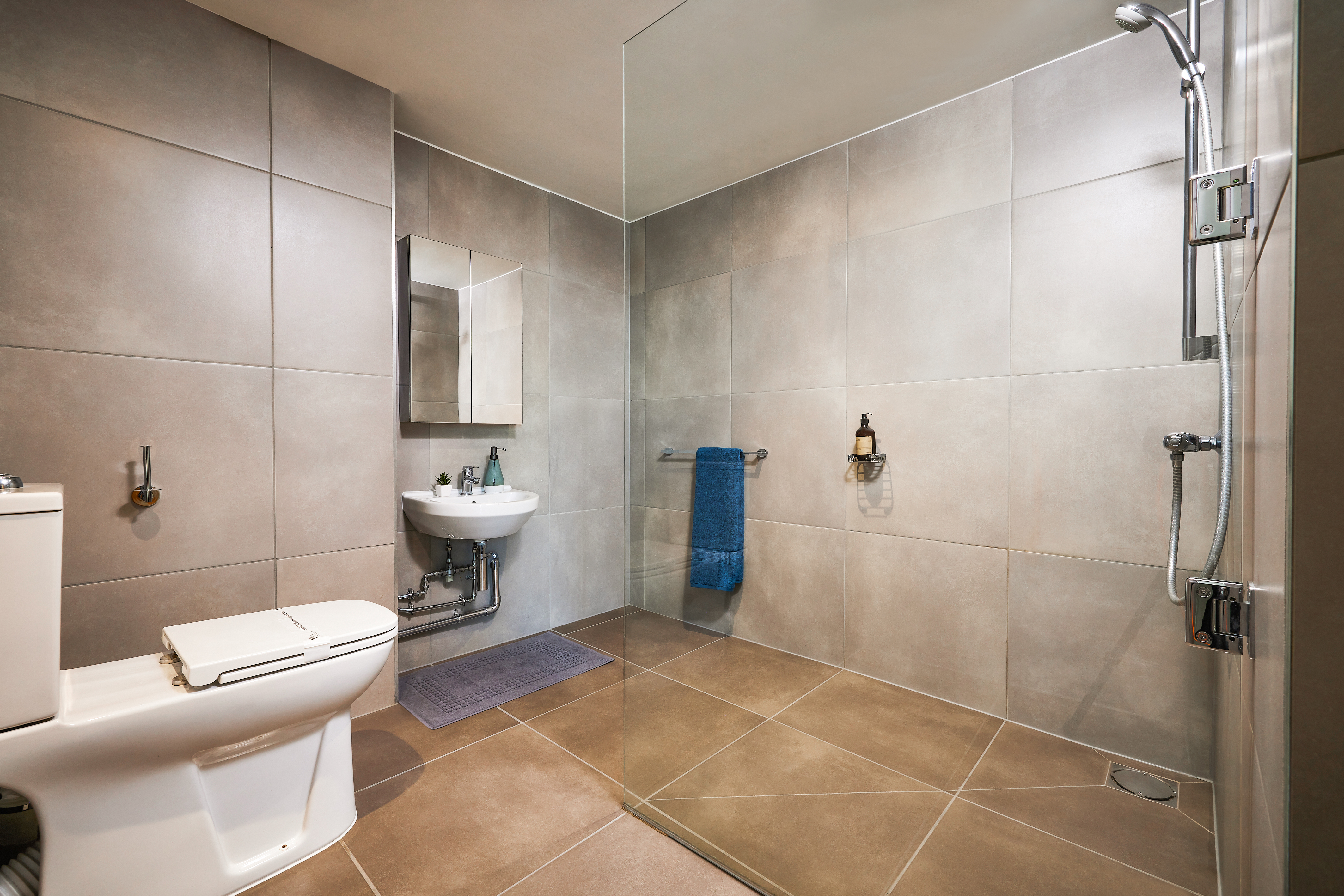 A modern bathroom featuring a sleek walk-in shower with a glass partition, a white toilet, and a wall-mounted sink with a mirror above. A blue towel hangs on the rail as neutral-toned tiles cover the walls and floor—a calming place to unwind.